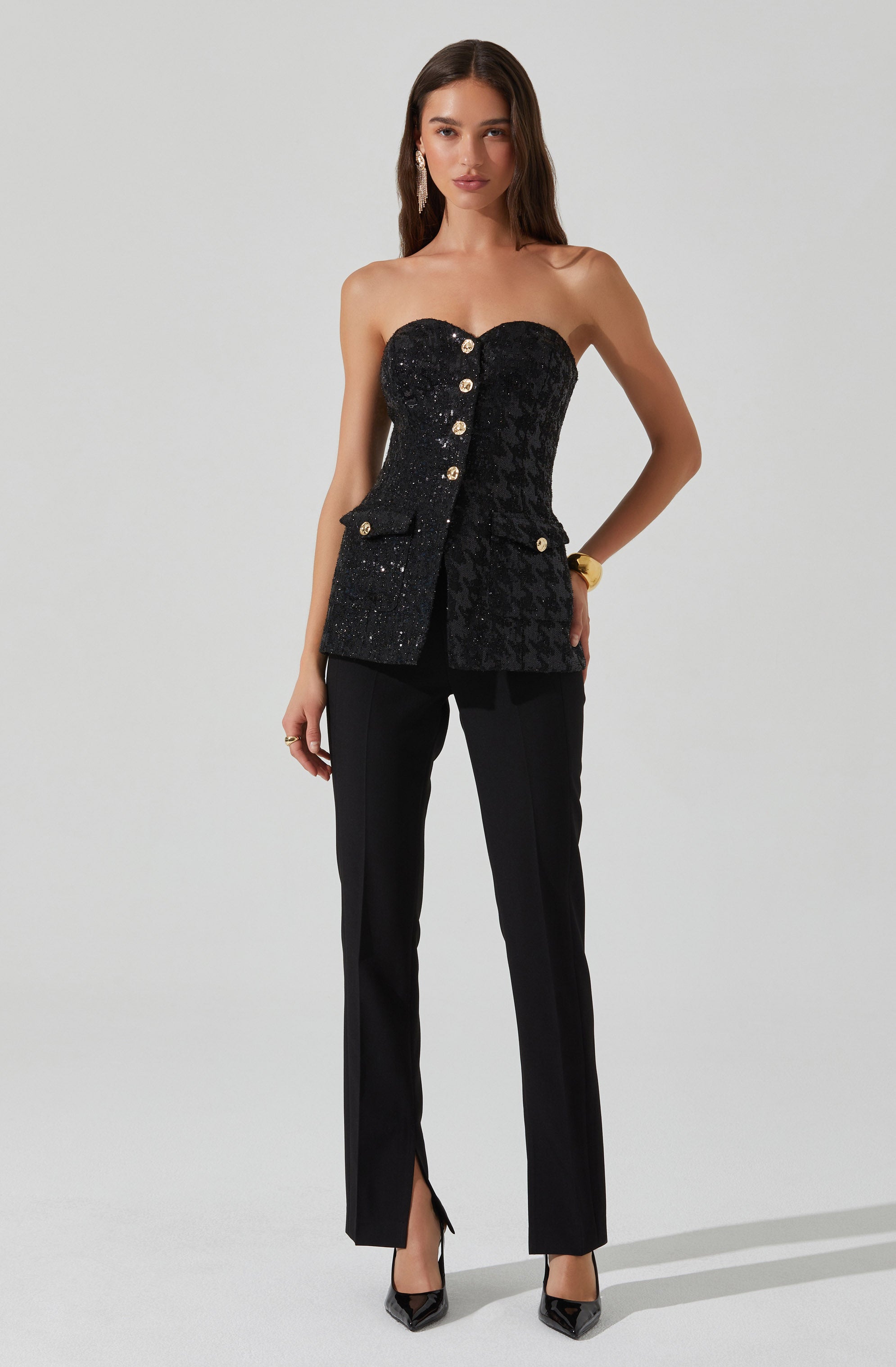 The woman is wearing a strapless black top that features a textured pattern and decorative gold buttons down the front, with a small pocket on the right side. She pairs the top with tailored black pants that have a slit at the hem. She is also wearing black pointed-toe heels and a gold bracelet on her right wrist.