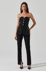 The woman is wearing a strapless black top that features a textured pattern and decorative gold buttons down the front, with a small pocket on the right side. She pairs the top with tailored black pants that have a slit at the hem. She is also wearing black pointed-toe heels and a gold bracelet on her right wrist.