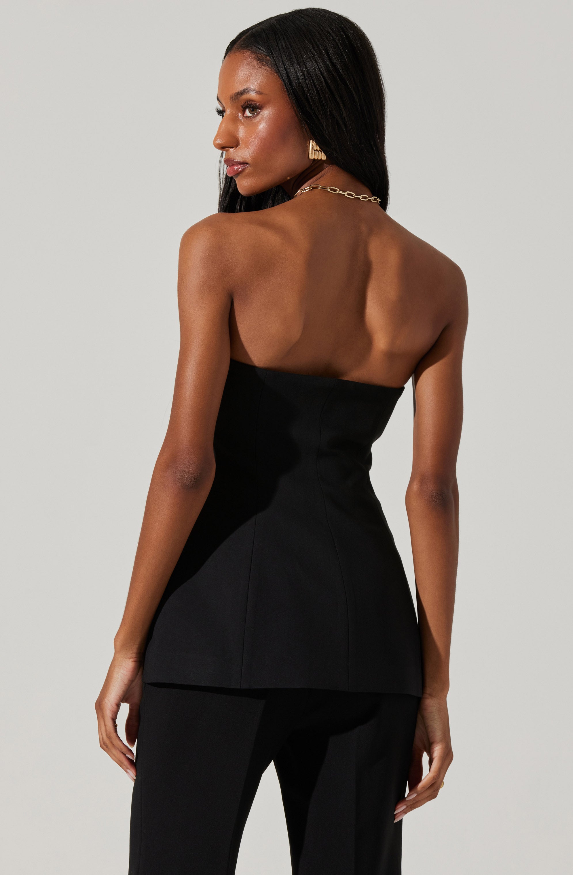 A woman is wearing a strapless black fitted top and matching black pants. The top is tailored and form-fitting, with a flared hemline at the waist. She accessorizes with gold earrings and a gold chain necklace.