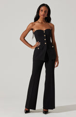 A woman is wearing a strapless black top with gold buttons down the front, paired with matching black wide-leg trousers. The top features a fitted bodice and decorative flap pockets on the hips with gold buttons. She accessorizes with a gold necklace and gold earrings, and is wearing black high heels.