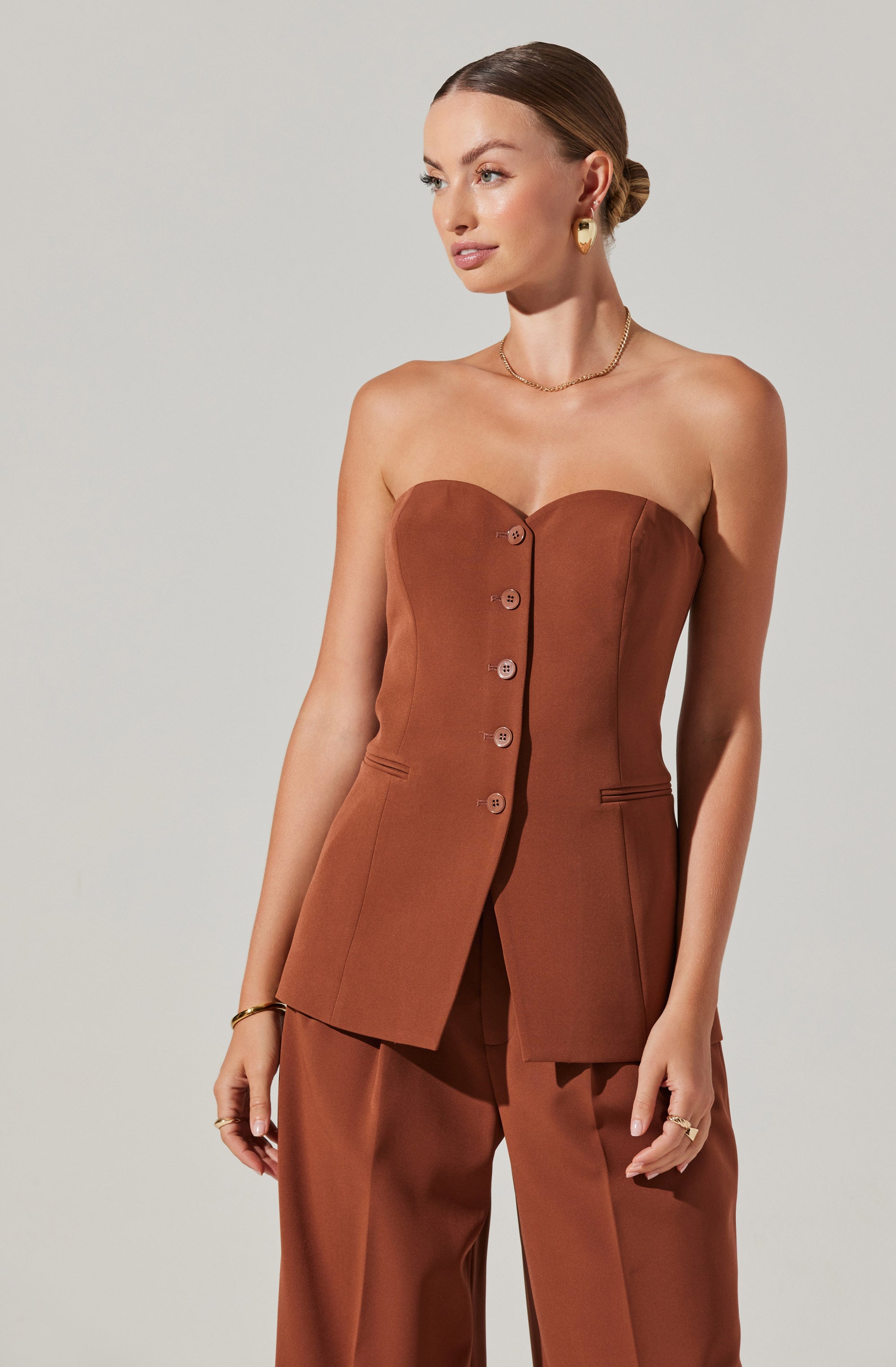 A woman is wearing a strapless, brown tailored suit consisting of a structured blazer with button closures down the front and matching high-waisted pants. She accessorizes with gold hoop earrings, a delicate gold necklace, and a bracelet. Her hair is styled in a low bun.