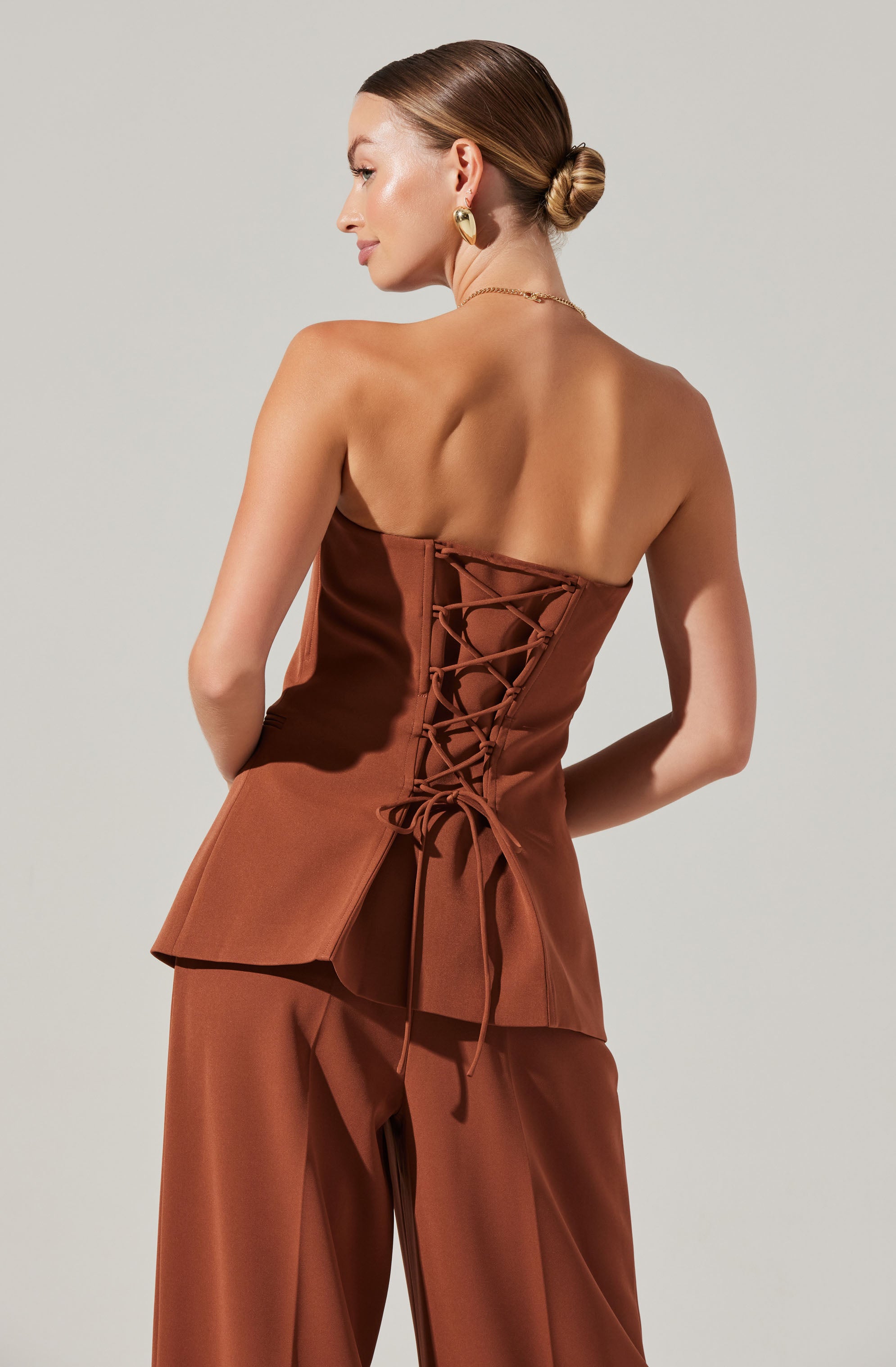 A woman is wearing a strapless, brown satin top with a lace-up back detail. The top features a peplum silhouette, and she pairs it with matching brown satin wide-leg pants.