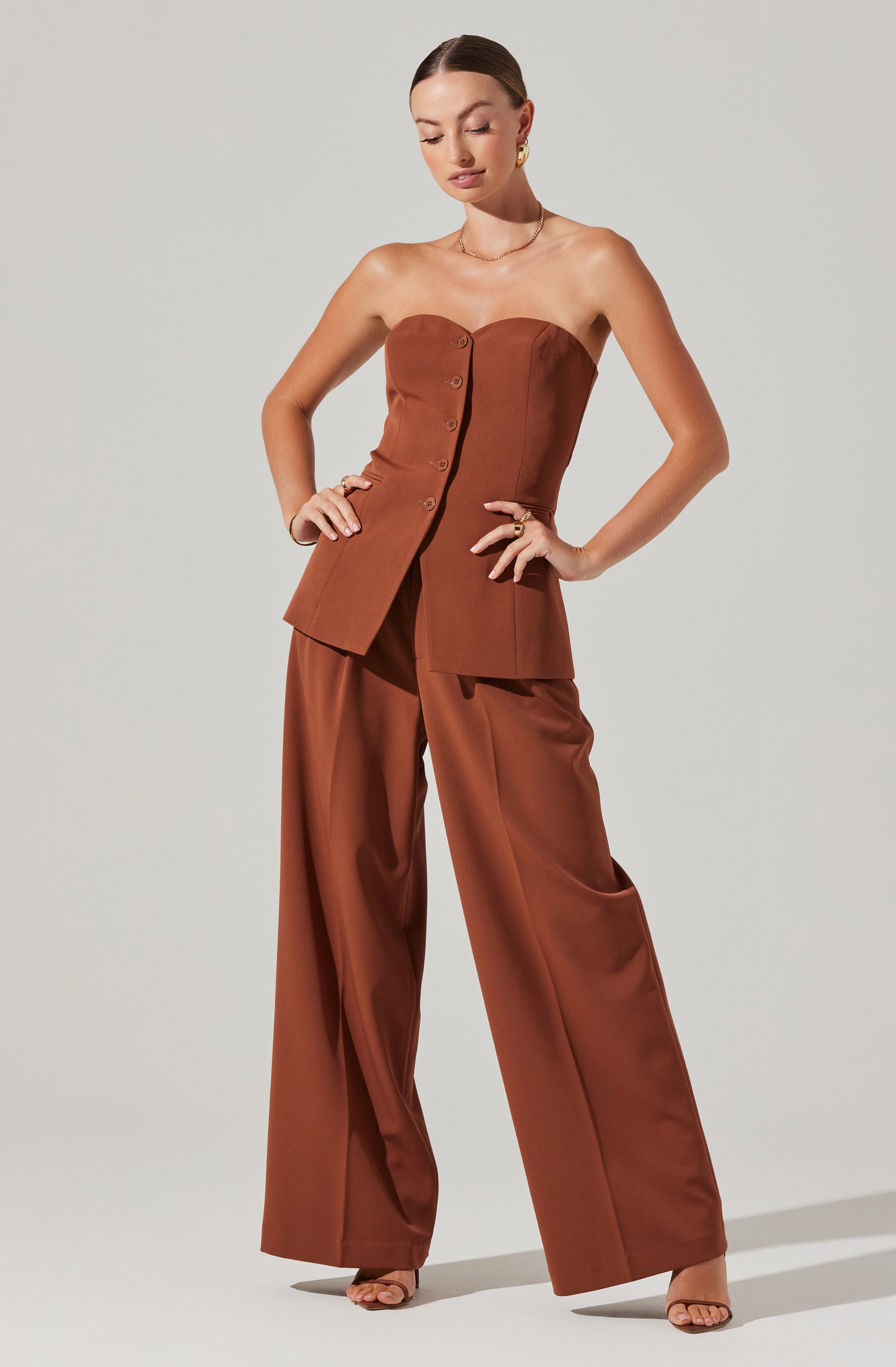 A woman is wearing a strapless, rust-colored bustier top with button details down the front, paired with matching wide-leg trousers. The outfit appears to be made of a smooth, satin-like fabric.
