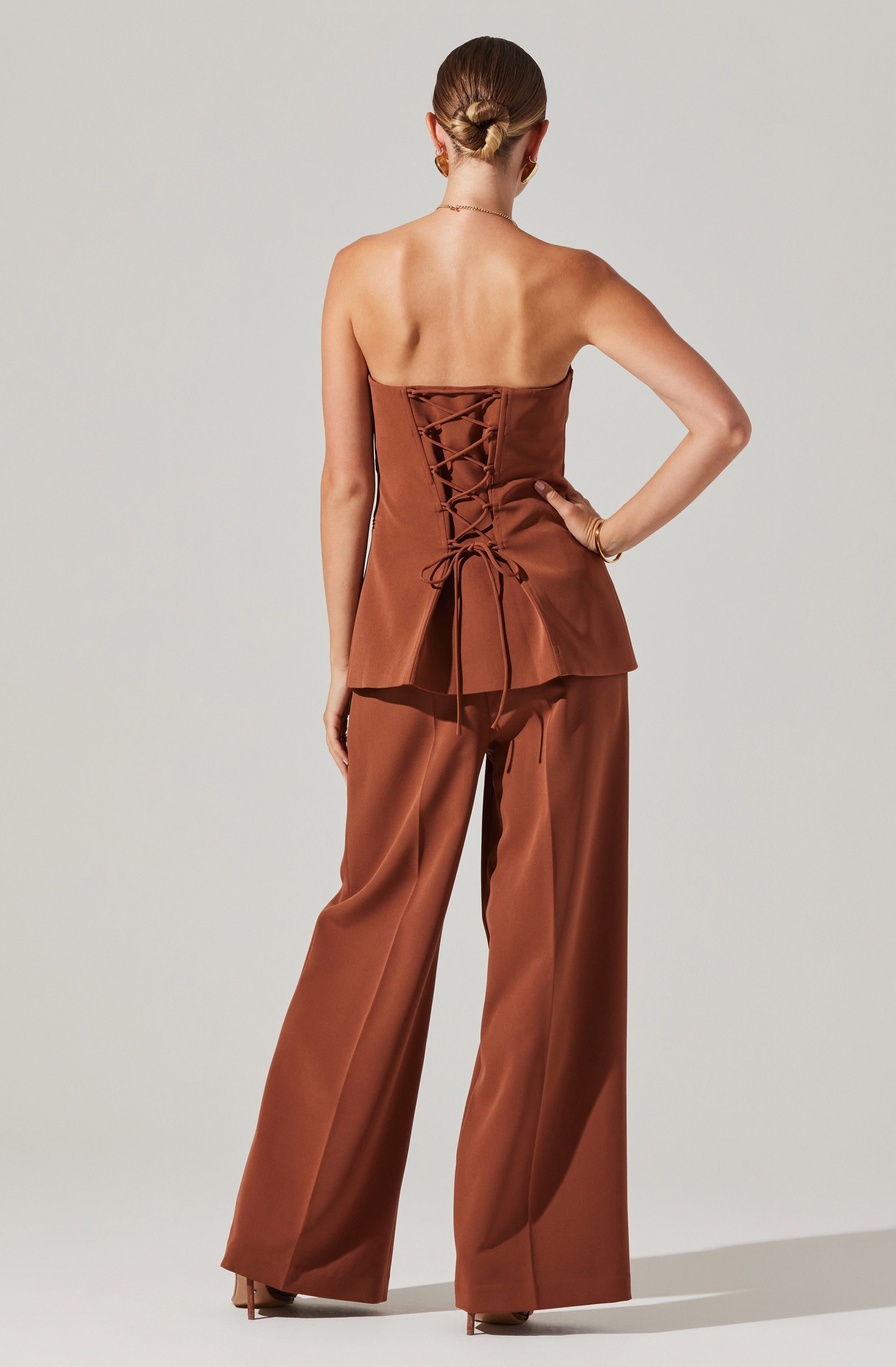 A woman is wearing a strapless brown satin top with a lace-up corset back and matching wide-leg pants.