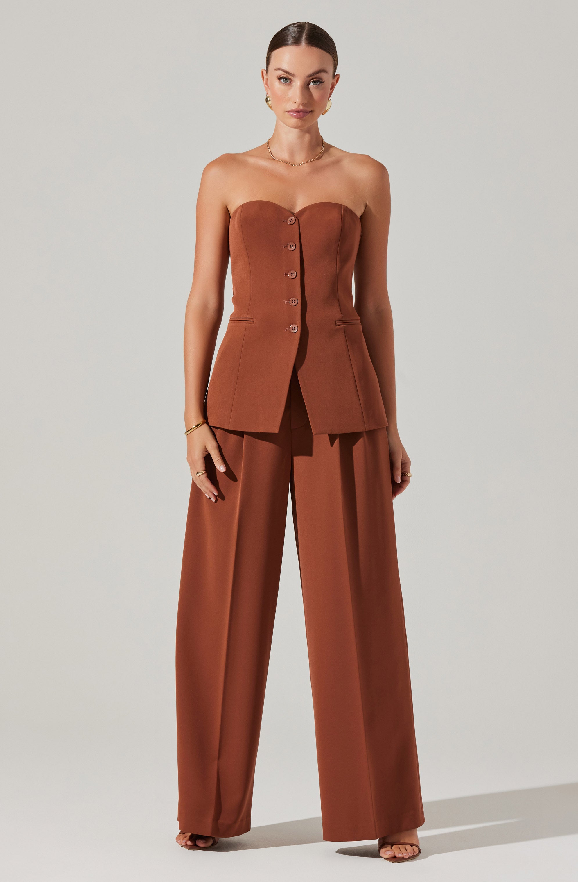 A woman is wearing a strapless brown sleeveless top with a sweetheart neckline and decorative buttons down the front. She pairs it with matching wide-leg brown trousers that have a high waist and pleats at the front.