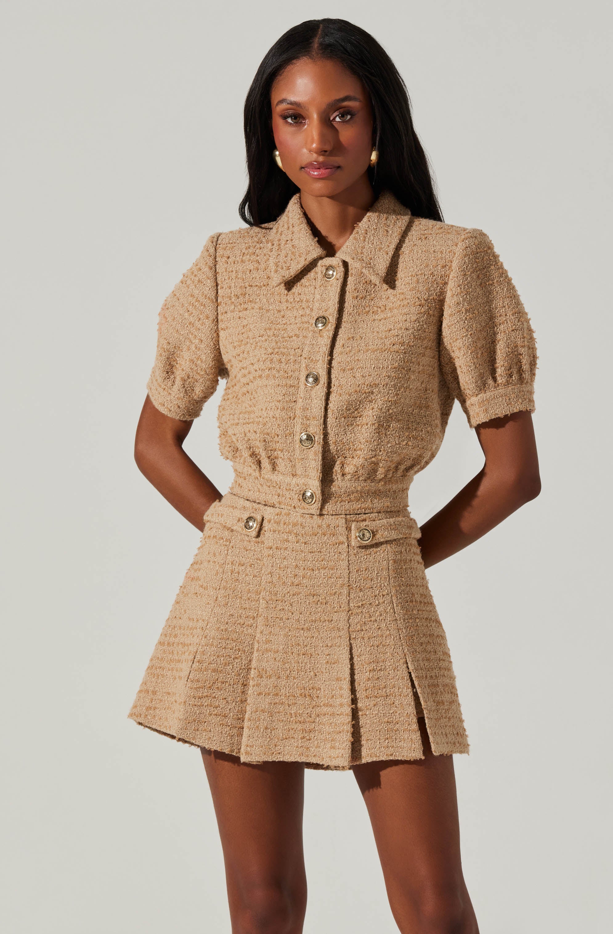 A woman is wearing a matching beige textured fabric outfit consisting of a short-sleeved button-up shirt with a collar and a high-waisted, pleated mini skirt. The shirt has puffed sleeves and gold buttons down the front. The skirt features a front slit and decorative buttons on the waistband.