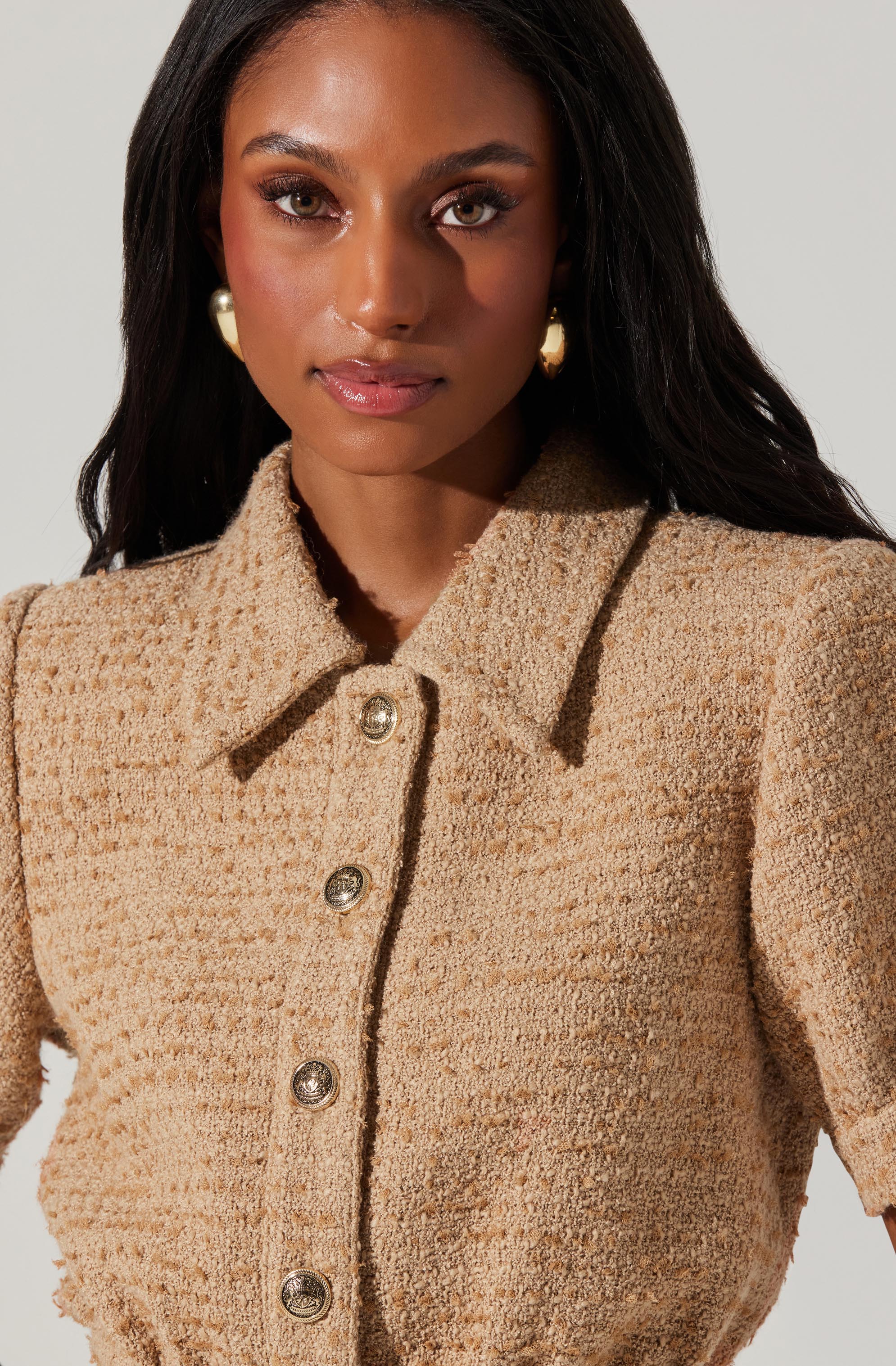 A woman is wearing a beige textured, woven jacket with a classic collar and large decorative buttons down the front. The jacket has puffed sleeves that end just above the elbows. She accessorizes with gold earrings.