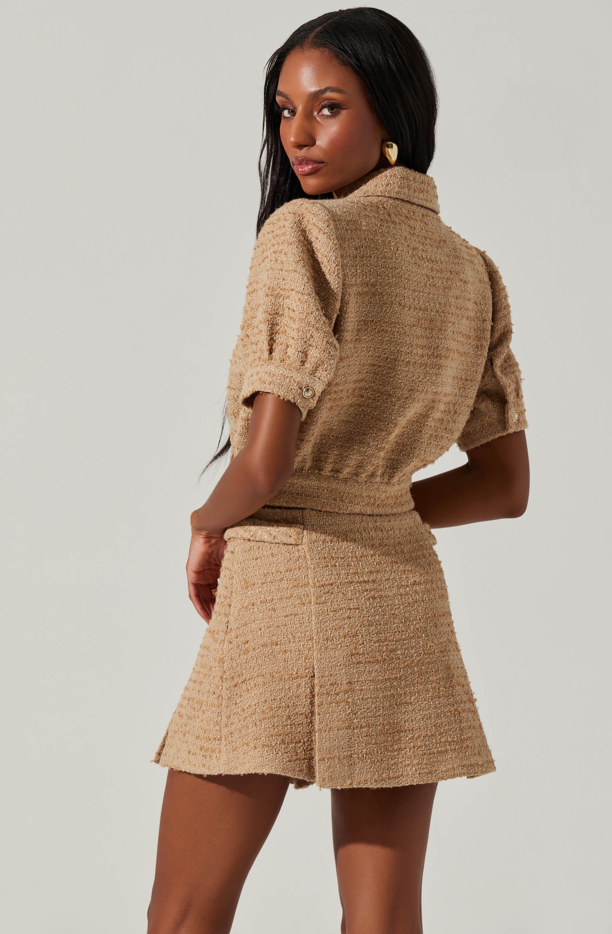 A woman is wearing a matching beige textured two-piece outfit comprising a short-sleeved, collared top with buttoned sleeve cuffs and a high-waisted, A-line mini skirt. The fabric appears to be a tweed or boucle material. She has long, dark hair and is accessorized with gold hoop earrings.