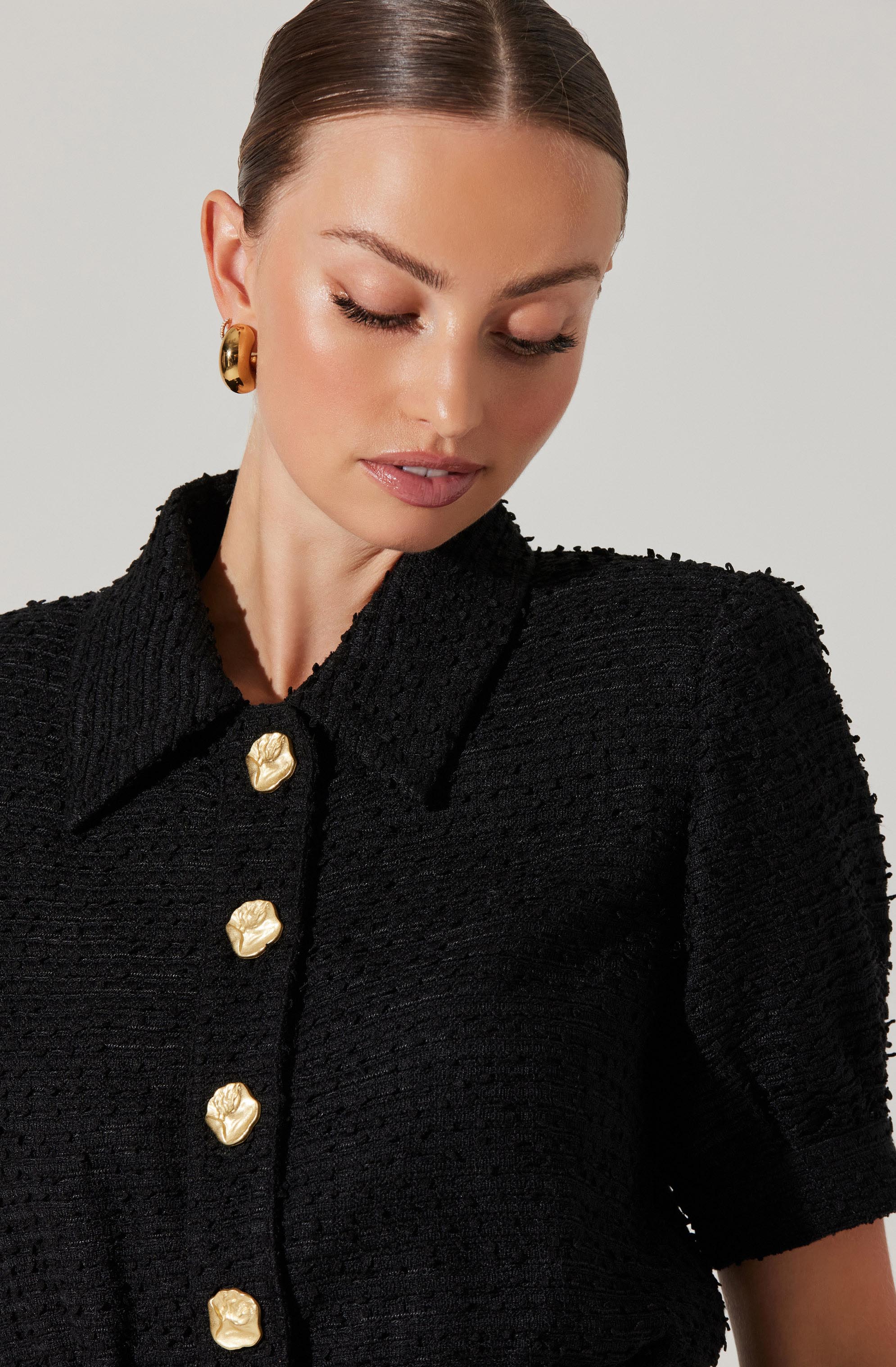 A woman is wearing a black textured short-sleeved jacket with a classic collar. The jacket features large, shiny gold buttons down the front, shaped like stylized flowers or shells. She is also wearing large, gold, twisted hoop earrings.