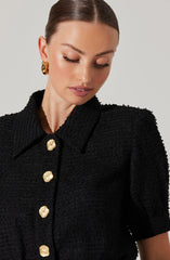 A woman is wearing a black textured short-sleeved jacket with a classic collar. The jacket features large, shiny gold buttons down the front, shaped like stylized flowers or shells. She is also wearing large, gold, twisted hoop earrings.