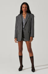 A woman is wearing a gray plaid oversized blazer with a notch lapel collar, a single button closure, and flap pockets. Underneath, she has a gray button-up shirt. She is also wearing black sheer knee-high socks and black pointed-toe high heels with decorative buckles.