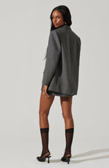 A woman is wearing an oversized gray plaid blazer with a classic collar and long sleeves. She pairs it with black sheer knee-high socks and black open-toe high heels. The blazer is the main focus of her outfit, giving a stylish contrast to her bare legs and shoes.