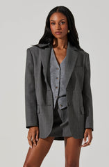A woman is dressed in a gray checked blazer with structured shoulders, paired with a matching gray cardigan and skirt underneath. The blazer has a classic notch lapel, and the cardigan features small buttons down the front. The skirt is matching in color and pattern, creating a coordinated, professional look.