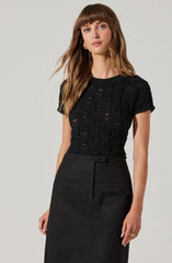 A woman is wearing a black short-sleeved knit top with a delicate, open weave pattern. She has paired it with a high-waisted black skirt that features a front zipper closure and a small belt loop.