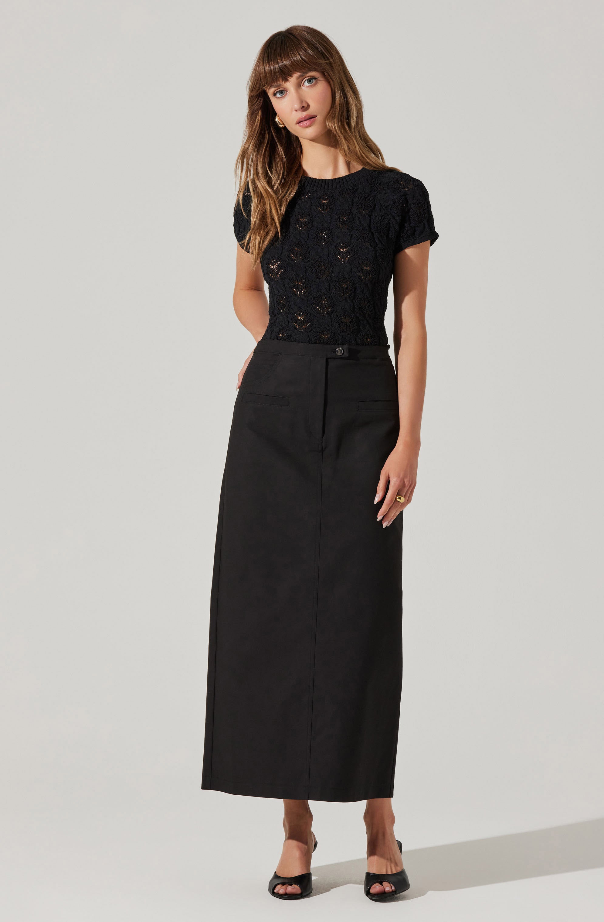 A woman is wearing a black short-sleeved, lace knit top with a round neckline, paired with a high-waisted, long black skirt that has a front slit and a single button closure at the waist. She completes her outfit with black open-toe heels.