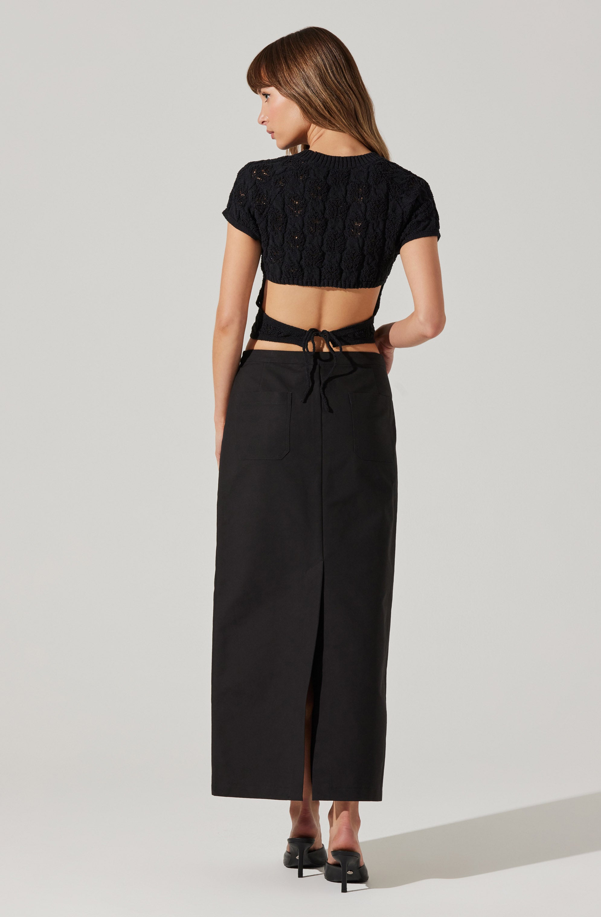 A woman is wearing a black, short-sleeved, lace crop top with a textured, open-weave pattern. She has tied a black ribbon around her waist, which is part of her long black skirt that reaches her ankles and features a front slit. She is also wearing black high-heeled shoes.