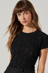 A woman is wearing a black lace short-sleeve top with a round neckline. The top features an intricate floral lace pattern that creates a semi-sheer effect. She pairs it with high-waisted black pants.