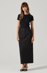 A woman is wearing a black, short-sleeved, lace top with a floral pattern. She pairs it with a high-waisted, long black skirt that reaches her ankles. She is also wearing black high-heeled sandals.