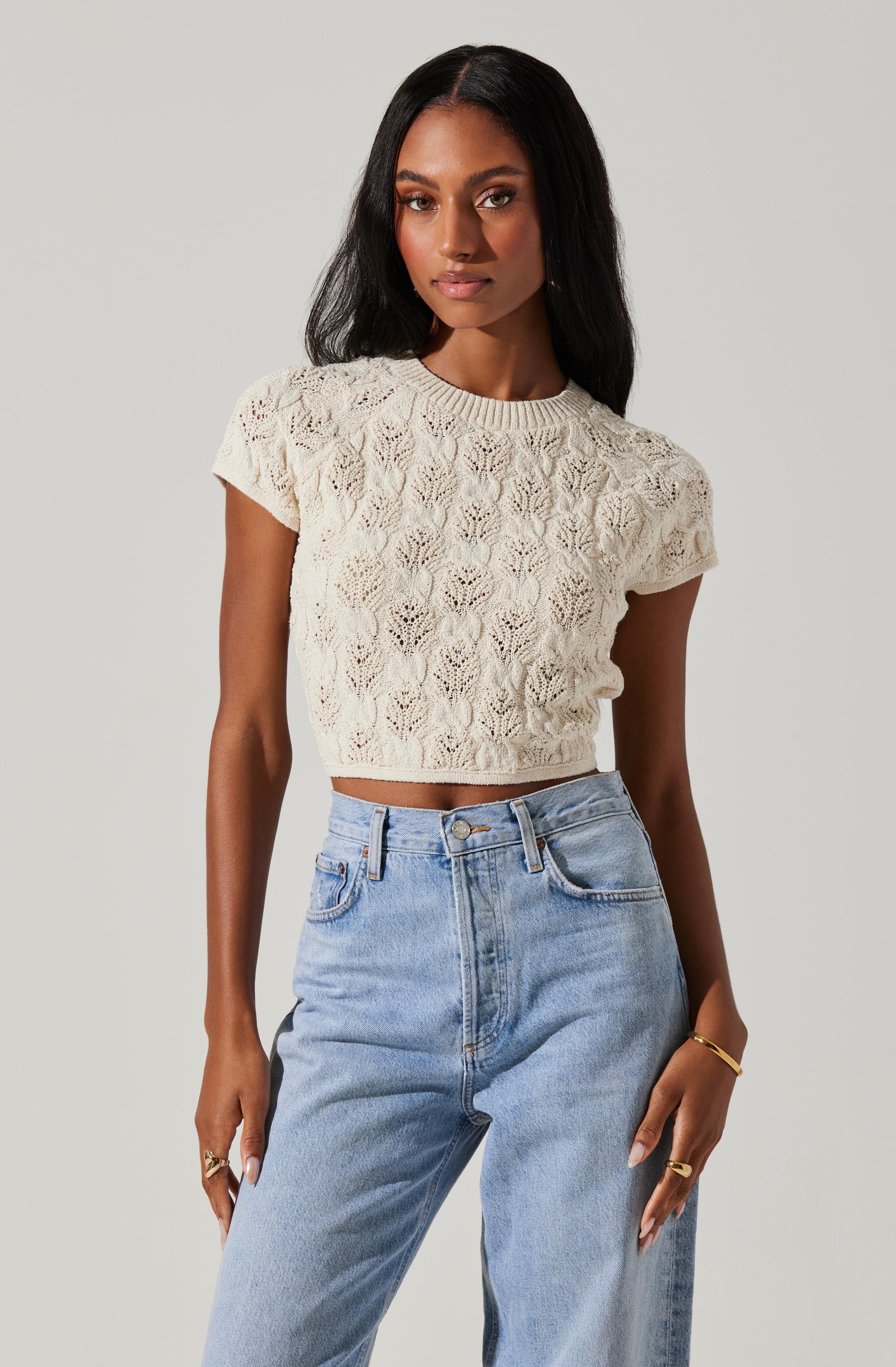 A woman is wearing a cream-colored, short-sleeved, cropped knit top with an intricate lace-like pattern. She pairs it with high-waisted, light-wash jeans that have a relaxed fit. She accessorizes with gold jewelry, including rings and a bracelet.