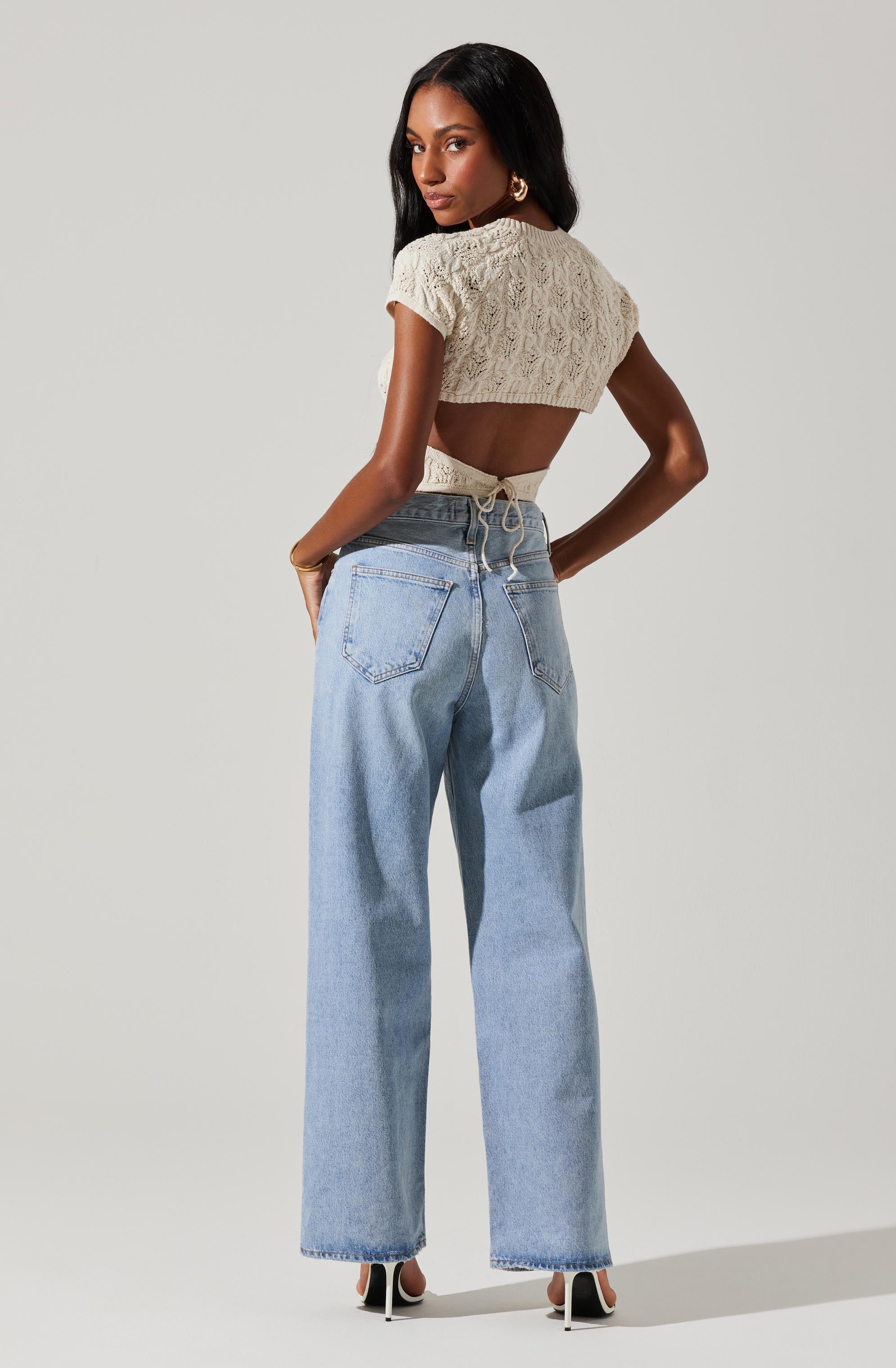 A woman is wearing a beige, short-sleeved, knitted crop top with a lace pattern, tied at the back with a bow. She is also wearing light blue high-waisted, wide-leg jeans with back pockets and white high-heeled shoes.