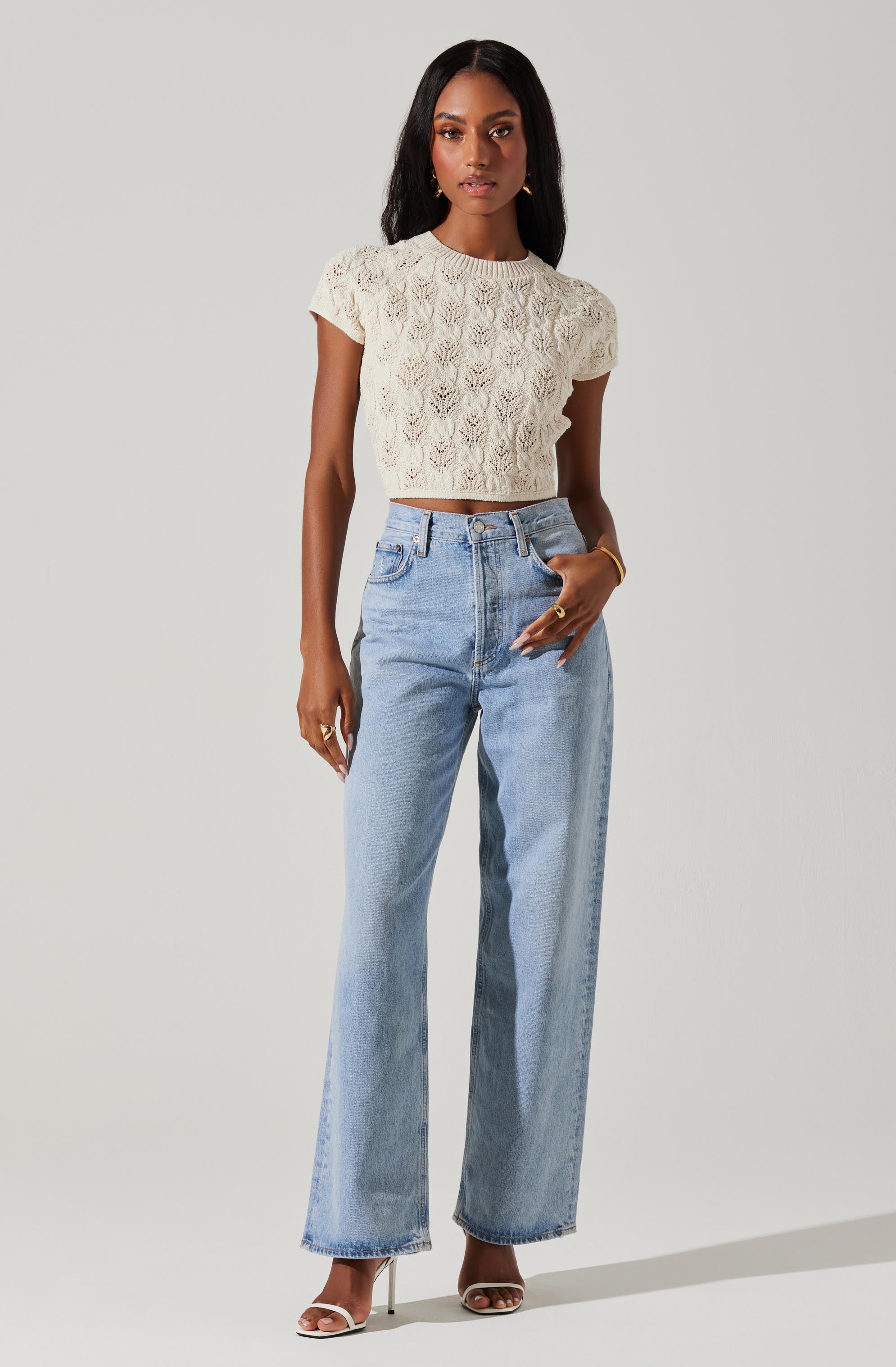 A woman wearing a short-sleeved, cream-colored crochet crop top with a textured, leafy pattern. She pairs it with high-waisted, light-wash wide-leg jeans that have a relaxed fit. She is accessorized with gold jewelry and is wearing white heeled sandals.