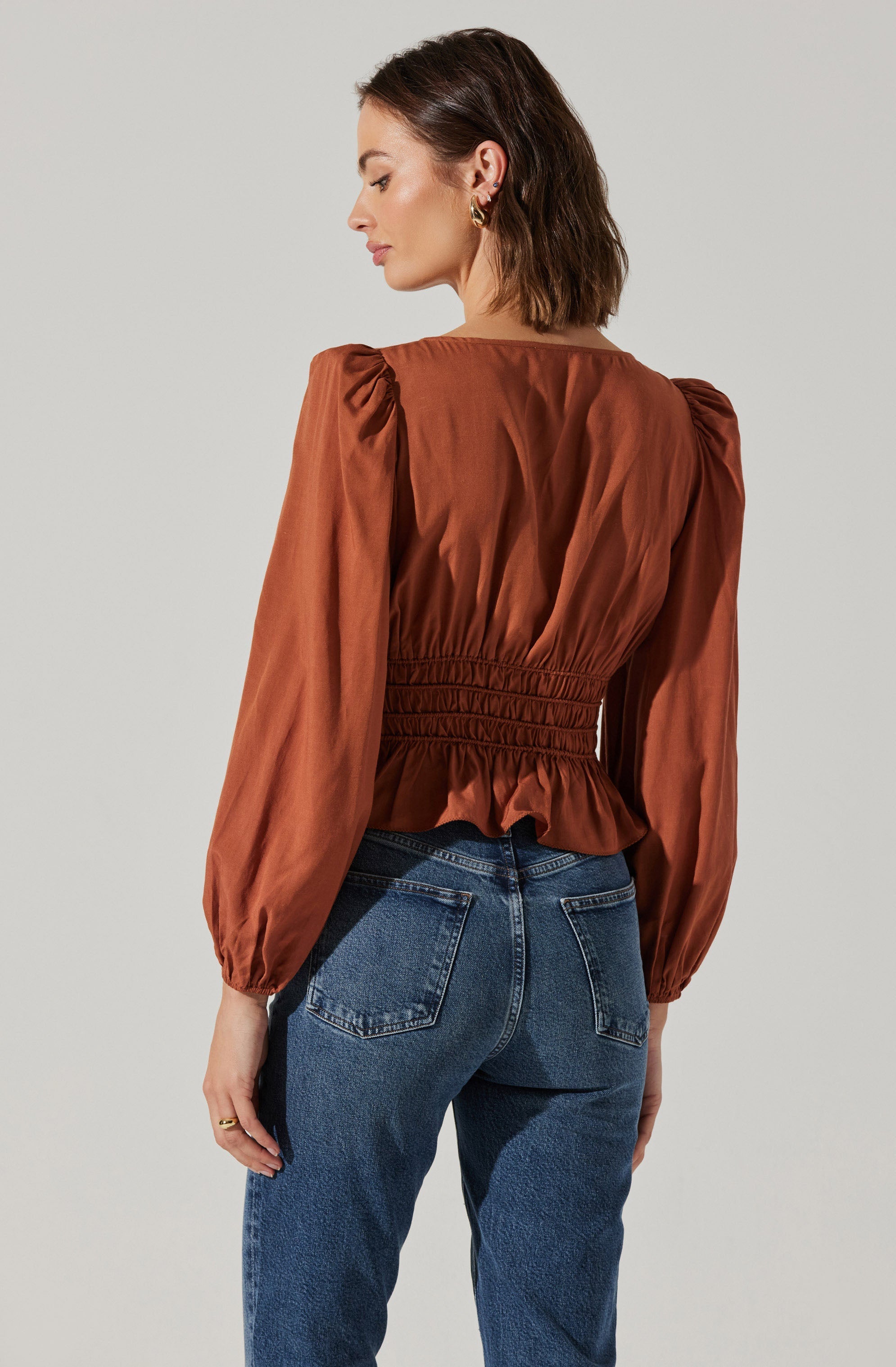 A woman is wearing a rust-colored blouse with long, puffed sleeves that gather at the wrists. The blouse has a fitted, smocked waist that creates a ruffled peplum effect at the hem. She is also wearing high-waisted blue denim jeans.