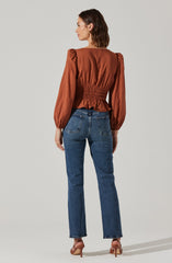 A woman is wearing a rust-colored blouse with long, puffed sleeves and a smocked waist. She pairs the blouse with high-waisted blue jeans that have visible back pockets. She is also wearing beige high-heeled shoes.