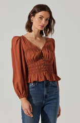A woman is wearing a rust-colored blouse with long puffed sleeves, a V-neckline, and smocked details around the waist and bust. The blouse has a slightly ruffled hem at the bottom. She is also wearing high-waisted blue jeans.