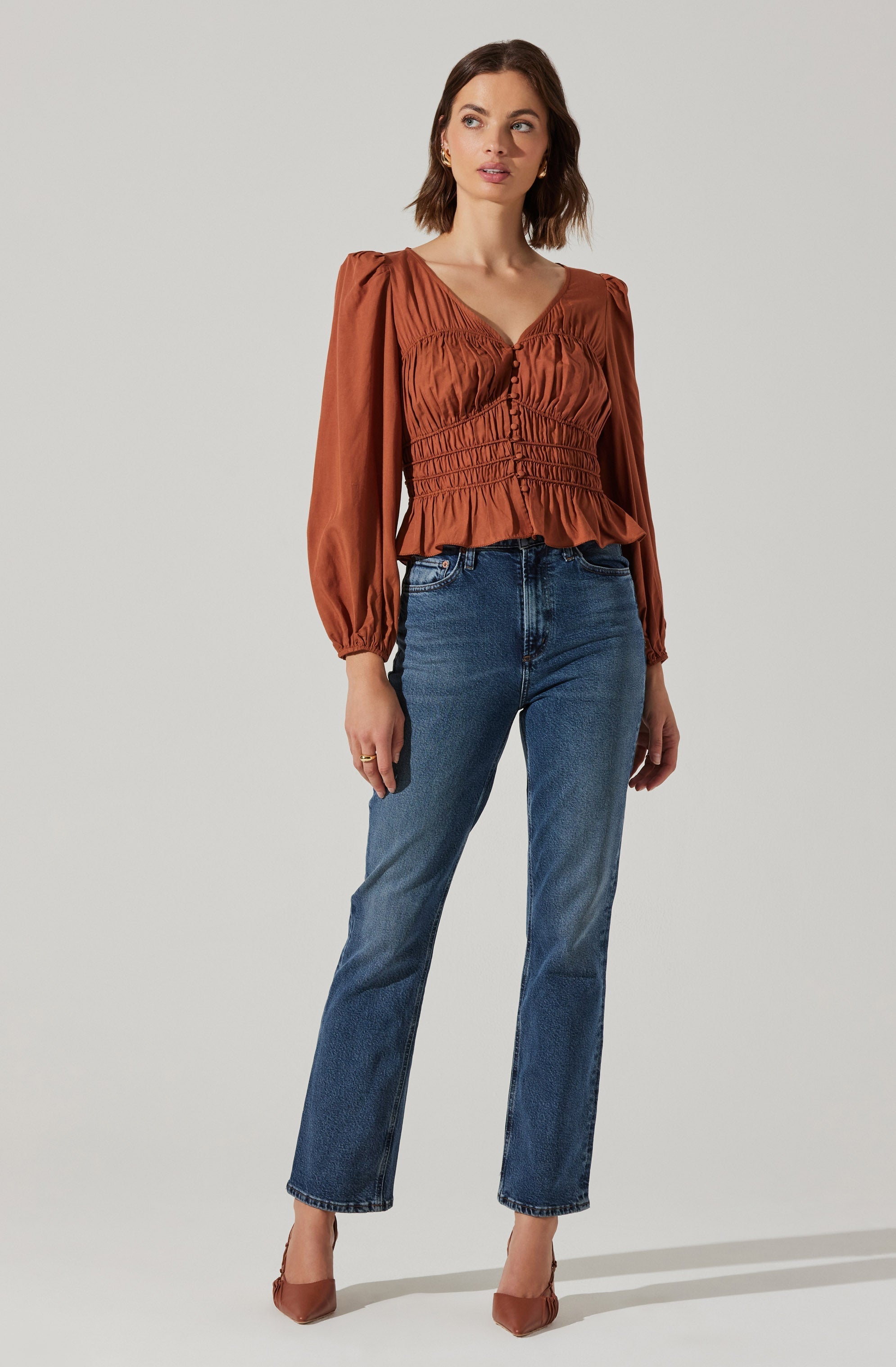 A woman is wearing a long-sleeved, rust-colored blouse with ruffles at the hem and a V-neckline. The blouse has voluminous sleeves gathered at the cuffs. She pairs it with high-waisted, straight-leg blue jeans and brown pointed-toe heels.