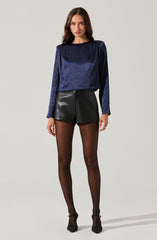 A woman is wearing a long-sleeved, dark blue, shimmery top paired with high-waisted black leather shorts, sheer black tights, and black high-heeled shoes.