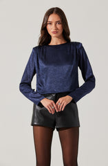 A woman is wearing a long-sleeved, dark blue, shimmery top with a round neckline. She is also wearing high-waisted black leather shorts and sheer black tights.