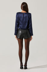 A woman is wearing a long-sleeved, shiny, navy blue blouse with a slight shimmer and a back slit. She paired it with high-waisted black leather shorts and sheer black tights. She is also wearing black high-heeled shoes.