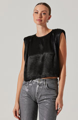 A woman is wearing a sleeveless black cropped top with a shiny, leather-like texture and slightly padded shoulders. She pairs it with high-waisted, metallic silver jeans.