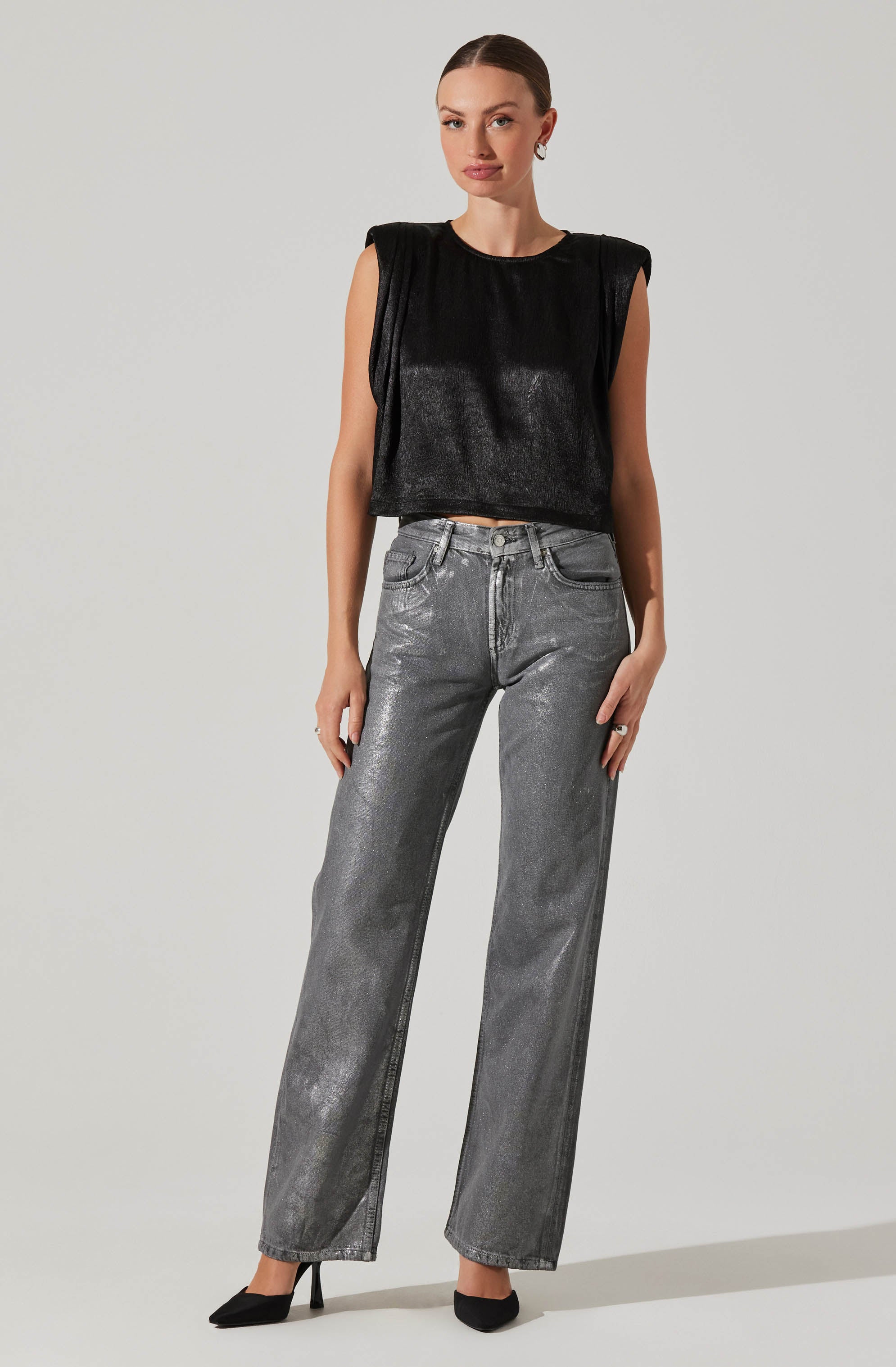 A woman is wearing a sleeveless black top with broad shoulder pads and shiny fabric, paired with high-waisted, straight-leg gray jeans that have a slightly metallic sheen. She completes her outfit with black pointed-toe high heels.