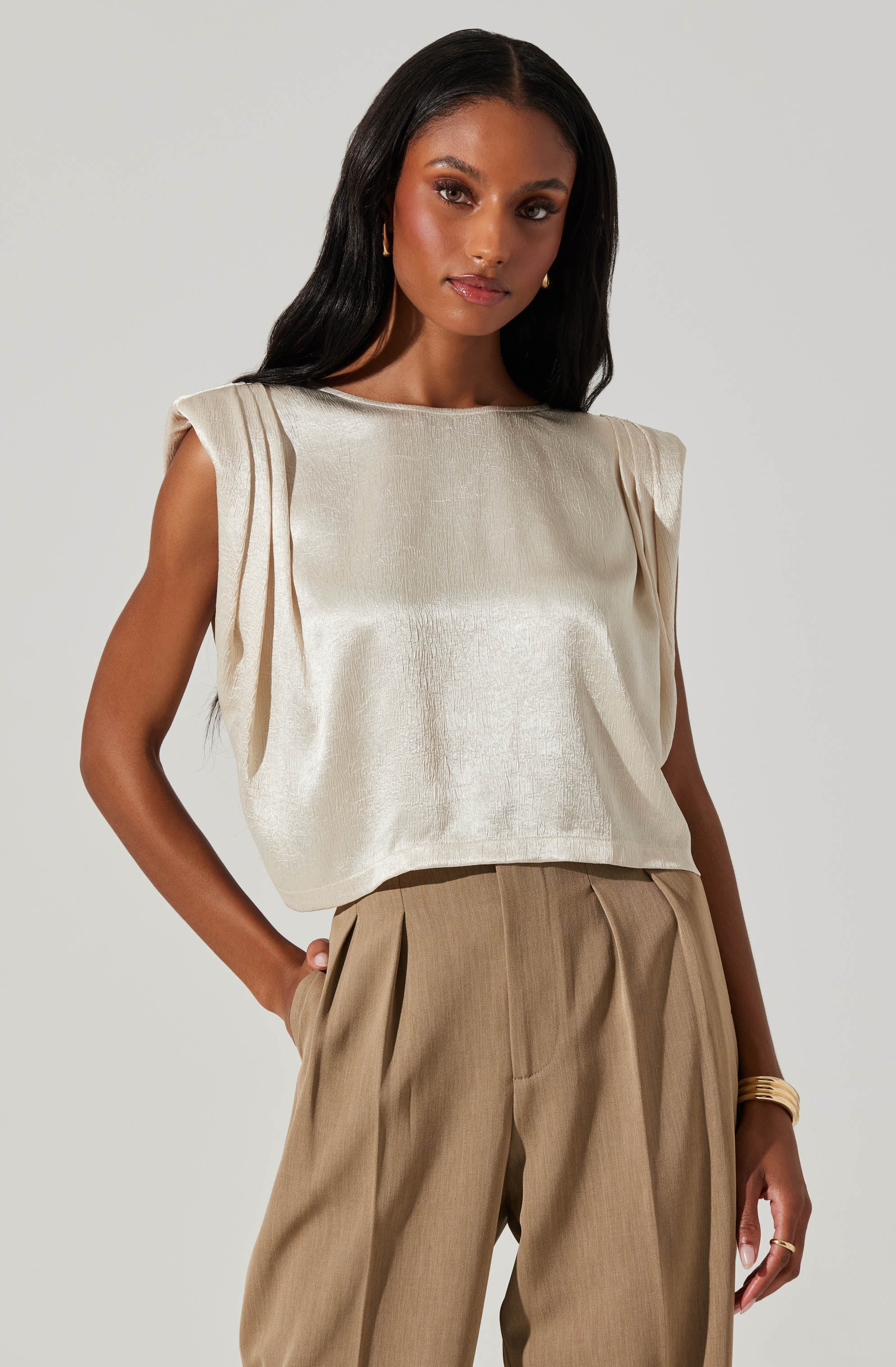 A woman is wearing a sleeveless, cream-colored, satin-like top with structured shoulder details. She pairs it with high-waisted, beige pleated trousers. She accessorizes with gold hoop earrings and a gold bracelet.