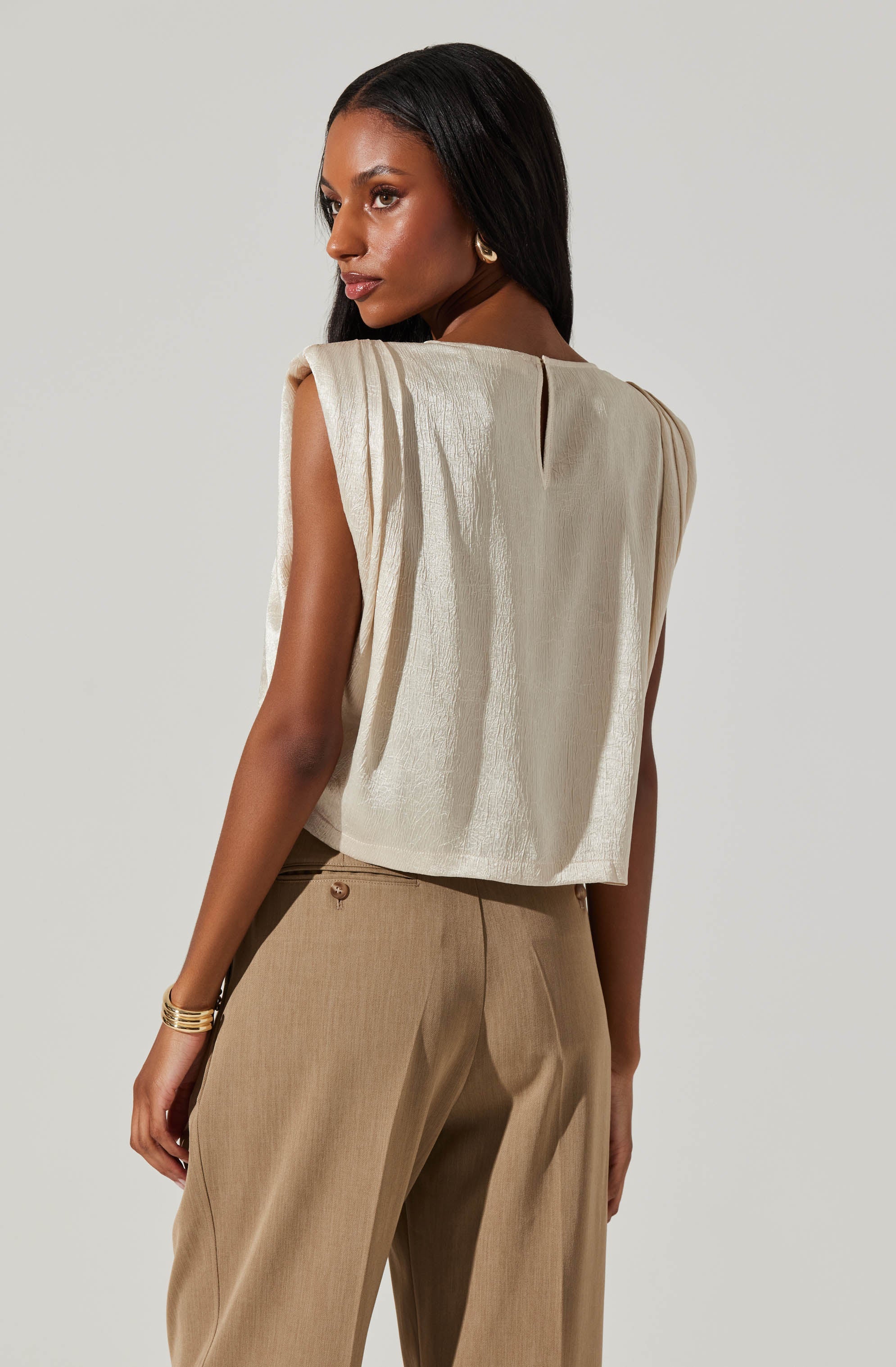 A woman is wearing a sleeveless, cream-colored top with a textured fabric and subtle pleats along the shoulders. The top has a slight boxy fit with a small back slit for ease of movement. She pairs it with high-waisted, tailored beige trousers that have a pleat at the front and a single button closure. She accessorizes with gold hoop earrings and a gold bracelet on her left wrist.