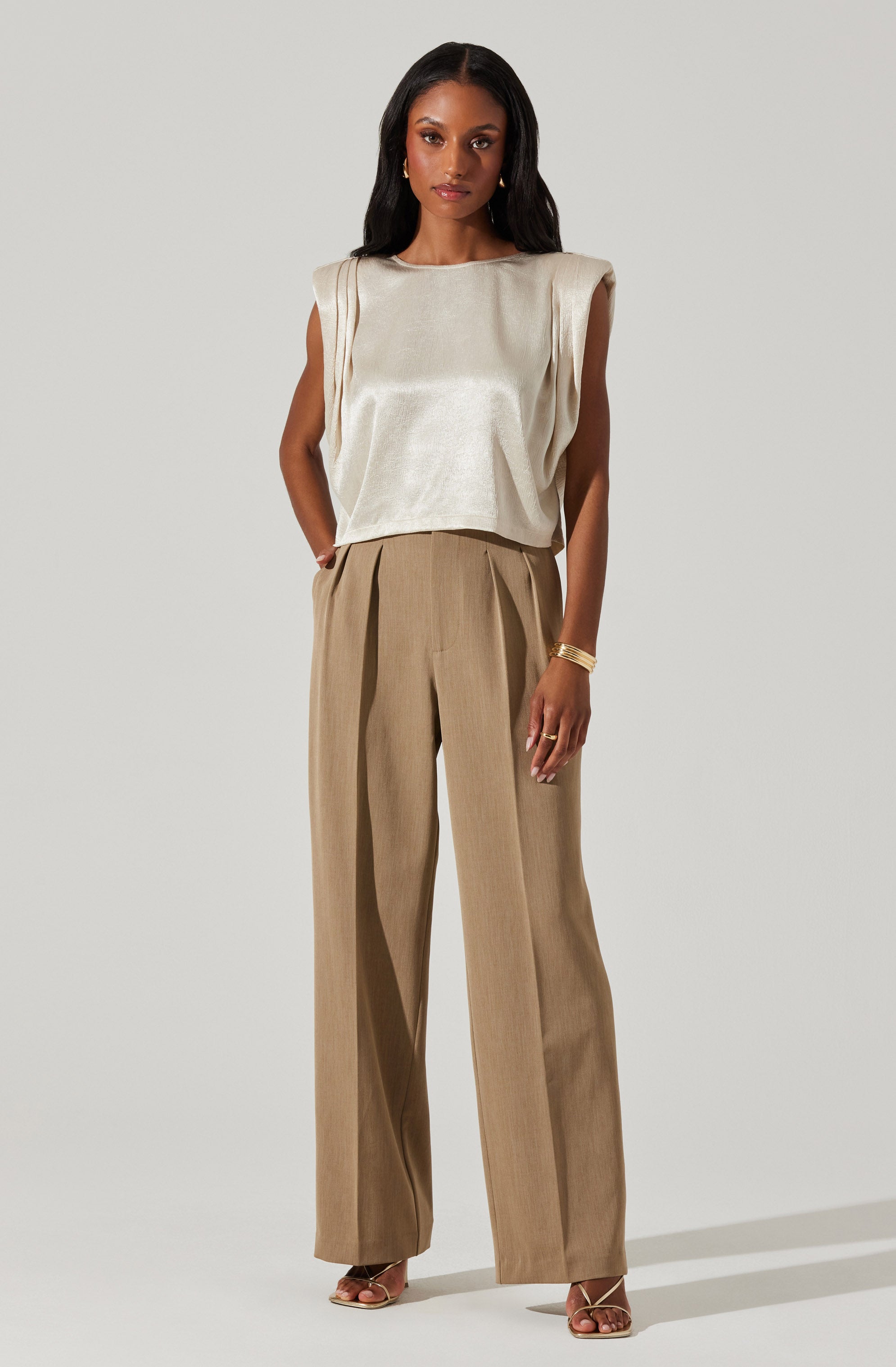 A woman is wearing a sleeveless, metallic beige top with padded shoulders and a relaxed fit. She pairs it with high-waisted, wide-leg tan trousers featuring pleats at the front. She accessorizes with gold jewelry, including a bracelet and earrings.