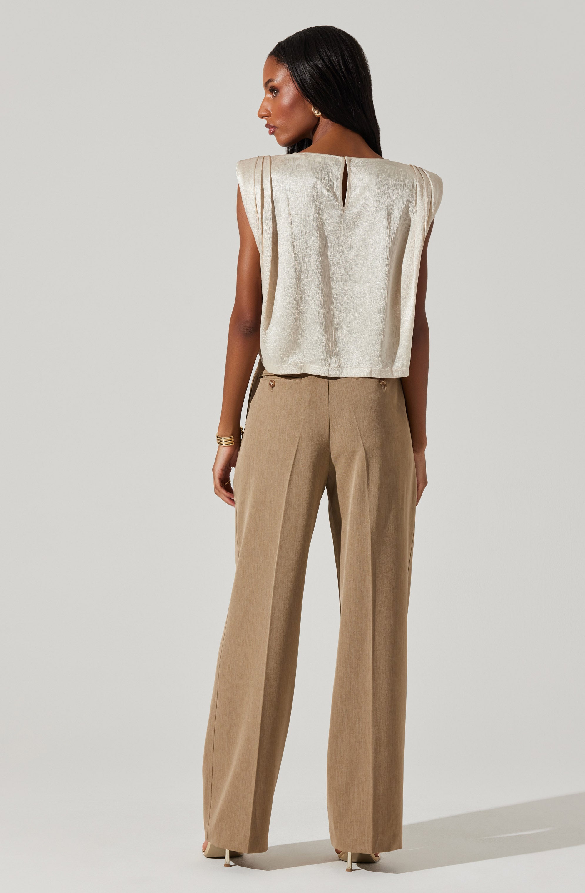 A woman is wearing a sleeveless, shimmery, cream-colored top with a small keyhole opening at the back neckline. She pairs it with high-waisted, beige wide-leg trousers that have a subtle pleat detail down the front.