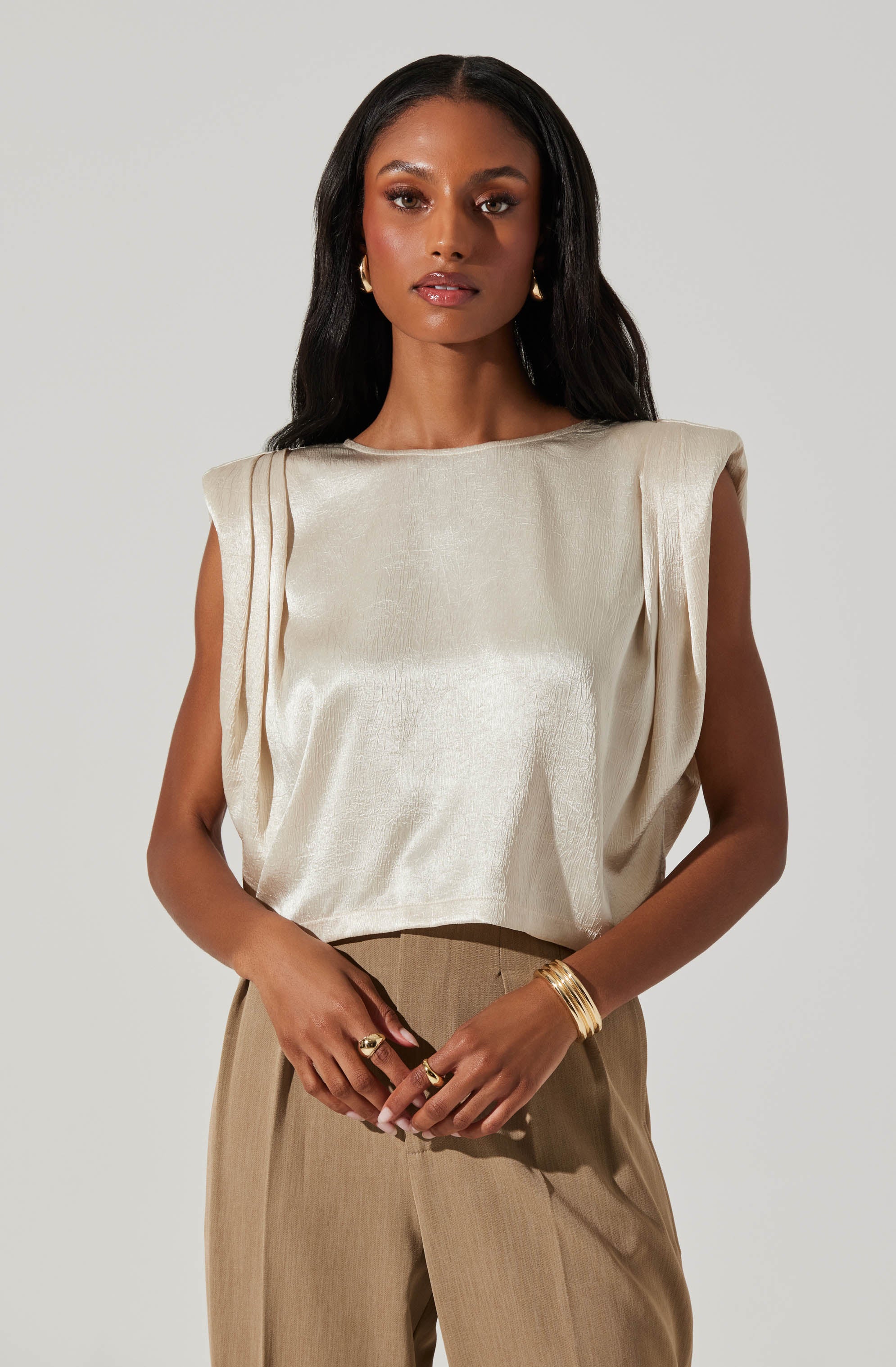 A woman is wearing a cream-colored satin sleeveless top with padded shoulders and a loose fit, paired with light brown high-waisted trousers. She accessorizes with gold hoop earrings, multiple gold bangles on her right wrist, and rings on both hands.
