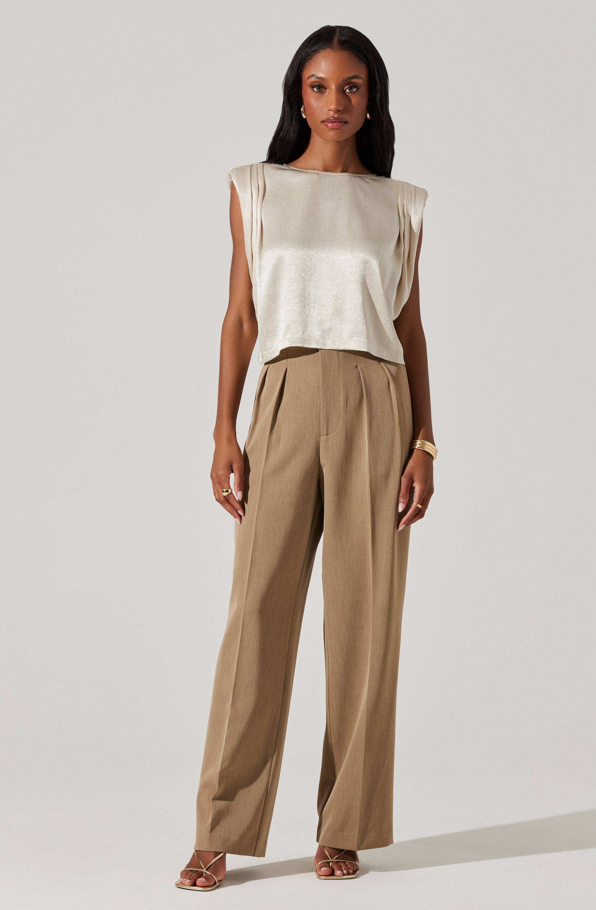 A woman is wearing a sleeveless, satin-like beige top with padded shoulders and a boxy cut. She pairs it with high-waisted, wide-leg tan trousers that have pleats at the front. She is also wearing open-toe heels and accessorized with gold jewelry, including earrings, rings, and a bracelet.