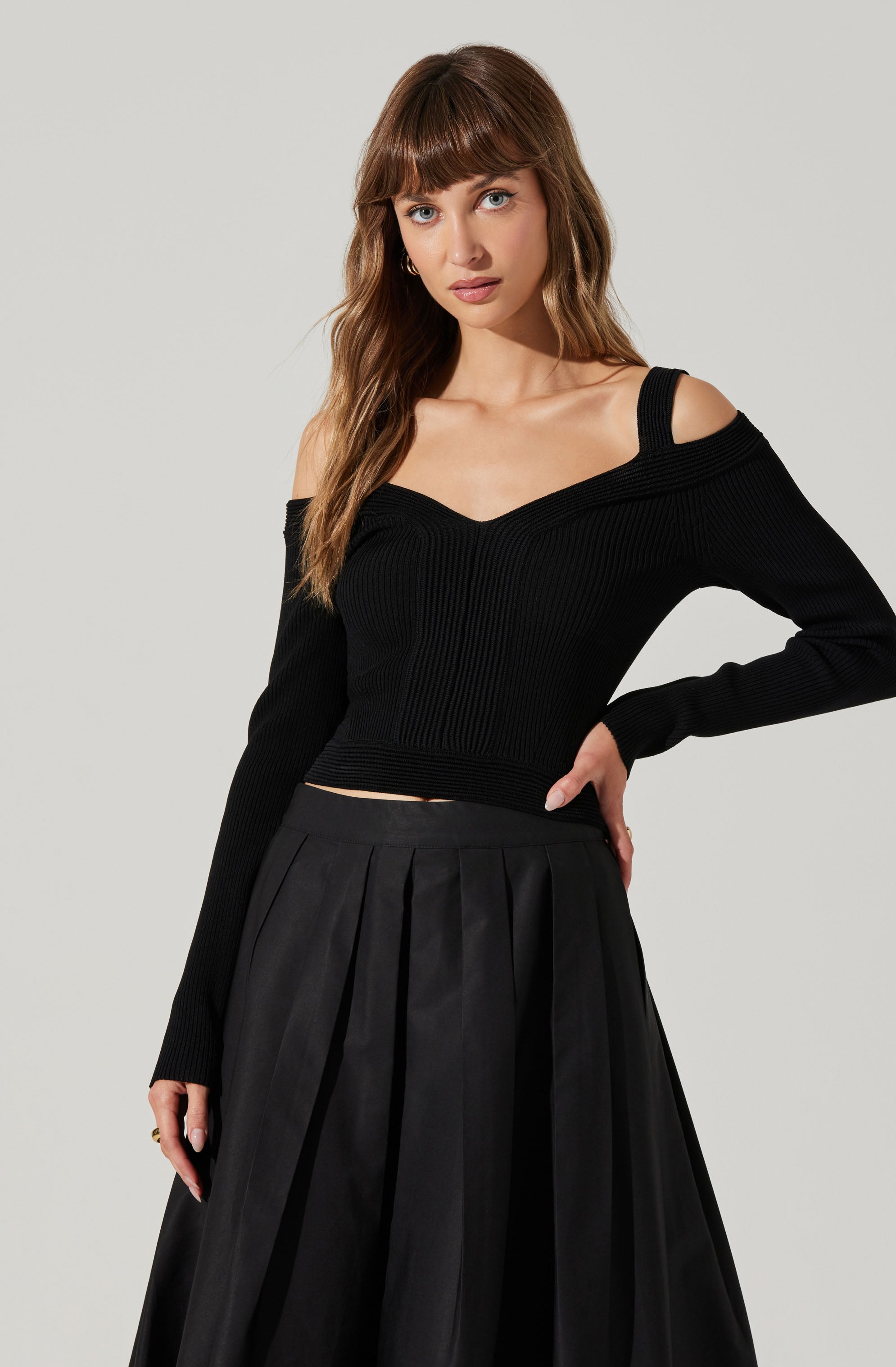 A young woman is wearing a black, fitted, long-sleeve sweater with a V-neck and off-the-shoulder design. The sweater has a ribbed texture and one strap over each shoulder. She pairs it with a high-waisted, black pleated skirt that falls below the knees.