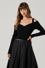 A young woman is wearing a black, fitted, long-sleeve sweater with a V-neck and off-the-shoulder design. The sweater has a ribbed texture and one strap over each shoulder. She pairs it with a high-waisted, black pleated skirt that falls below the knees.