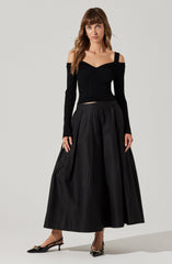 A woman is wearing a fitted, long-sleeved black sweater with a V-neckline and shoulder cut-outs. She pairs it with a high-waisted, full-length black skirt with pleats. She completes the outfit with black pointed-toe heels featuring a decorative chain detail across the front.