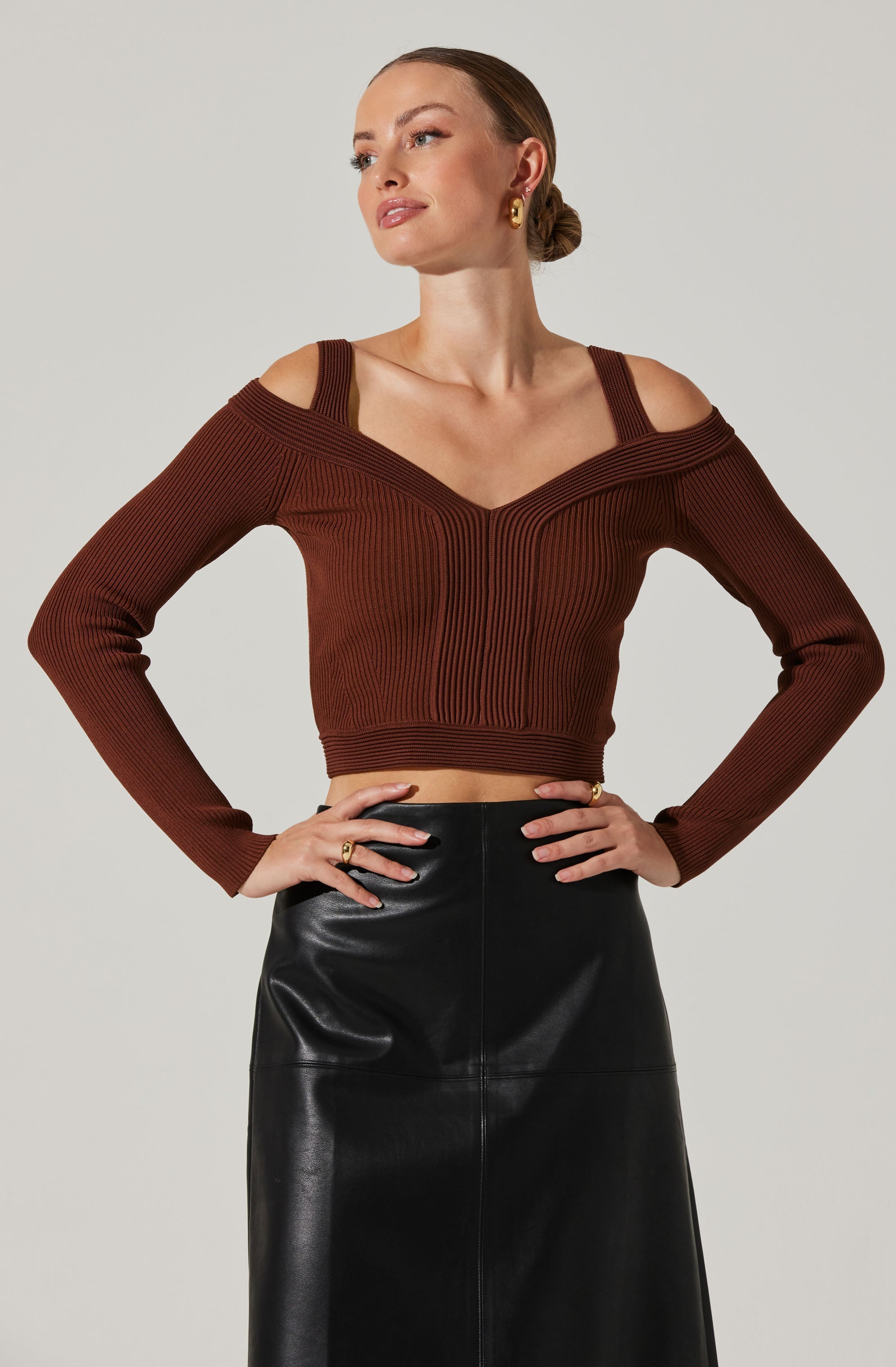 A woman is wearing a long-sleeve, brown, ribbed knit top with a unique design featuring cut-out shoulders and a deep V neckline that extends to the shoulders. She pairs it with a high-waisted, black leather skirt. She accessorizes with gold hoop earrings and rings.