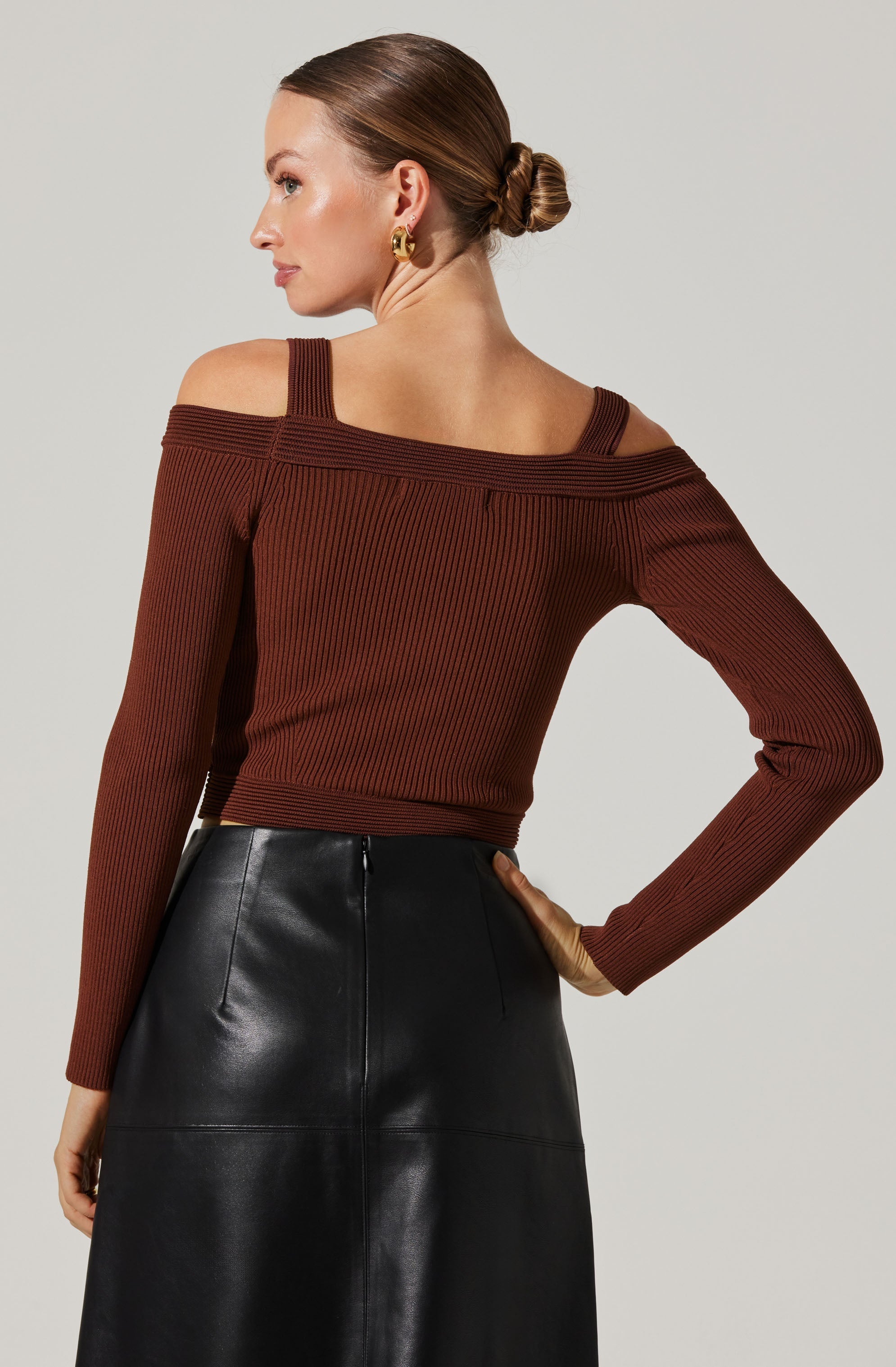 A woman is wearing a long-sleeve, off-the-shoulder sweater in a rich brown color with a ribbed texture and wide shoulder straps. She pairs it with a high-waisted, black leather skirt that has visible seams and a sleek, shiny finish.