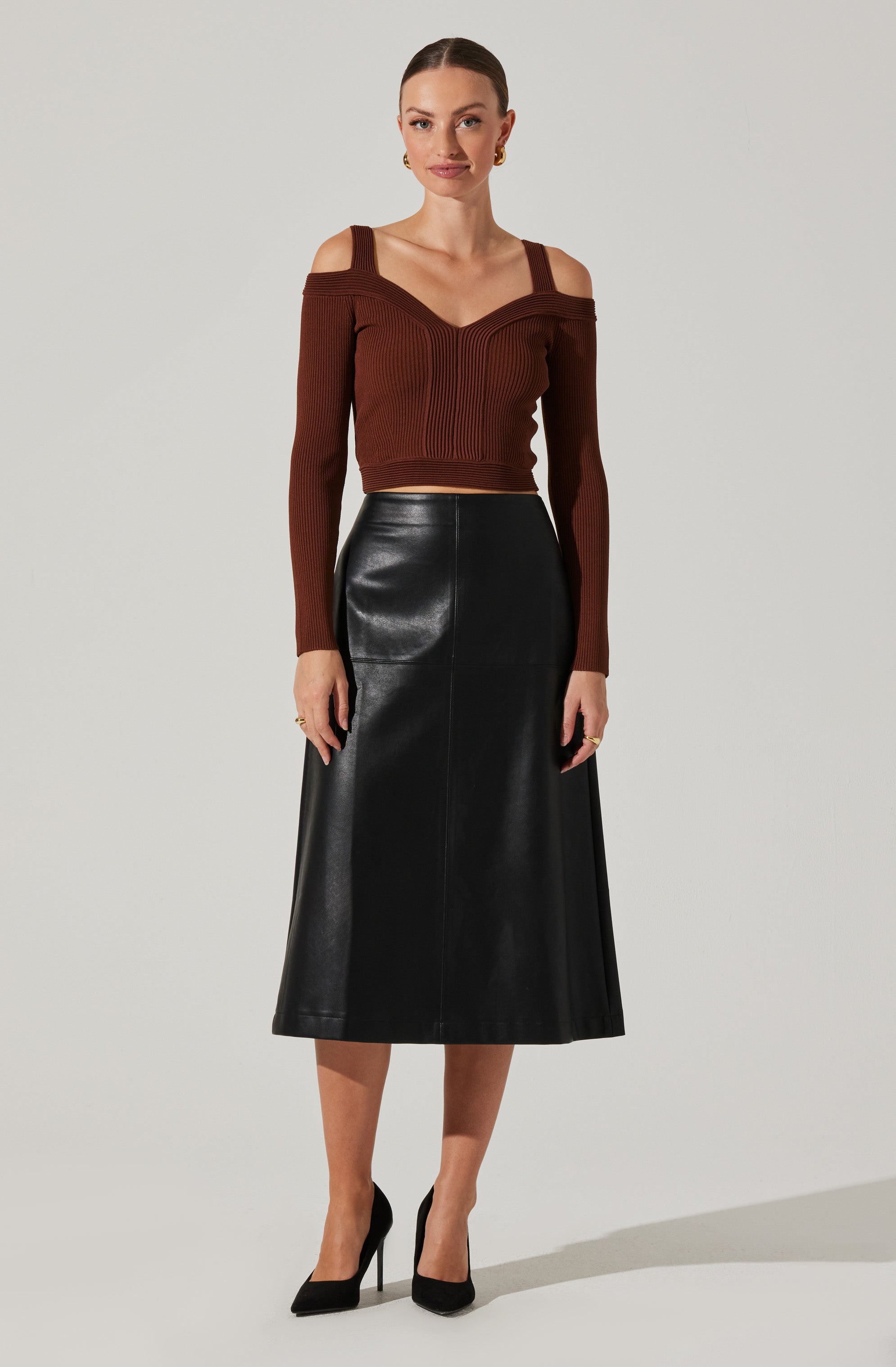 A woman is wearing a brown, off-the-shoulder, ribbed sweater with wide shoulder straps. She pairs it with a high-waisted, long black leather skirt that falls below her knees. She is also wearing black high-heeled shoes.