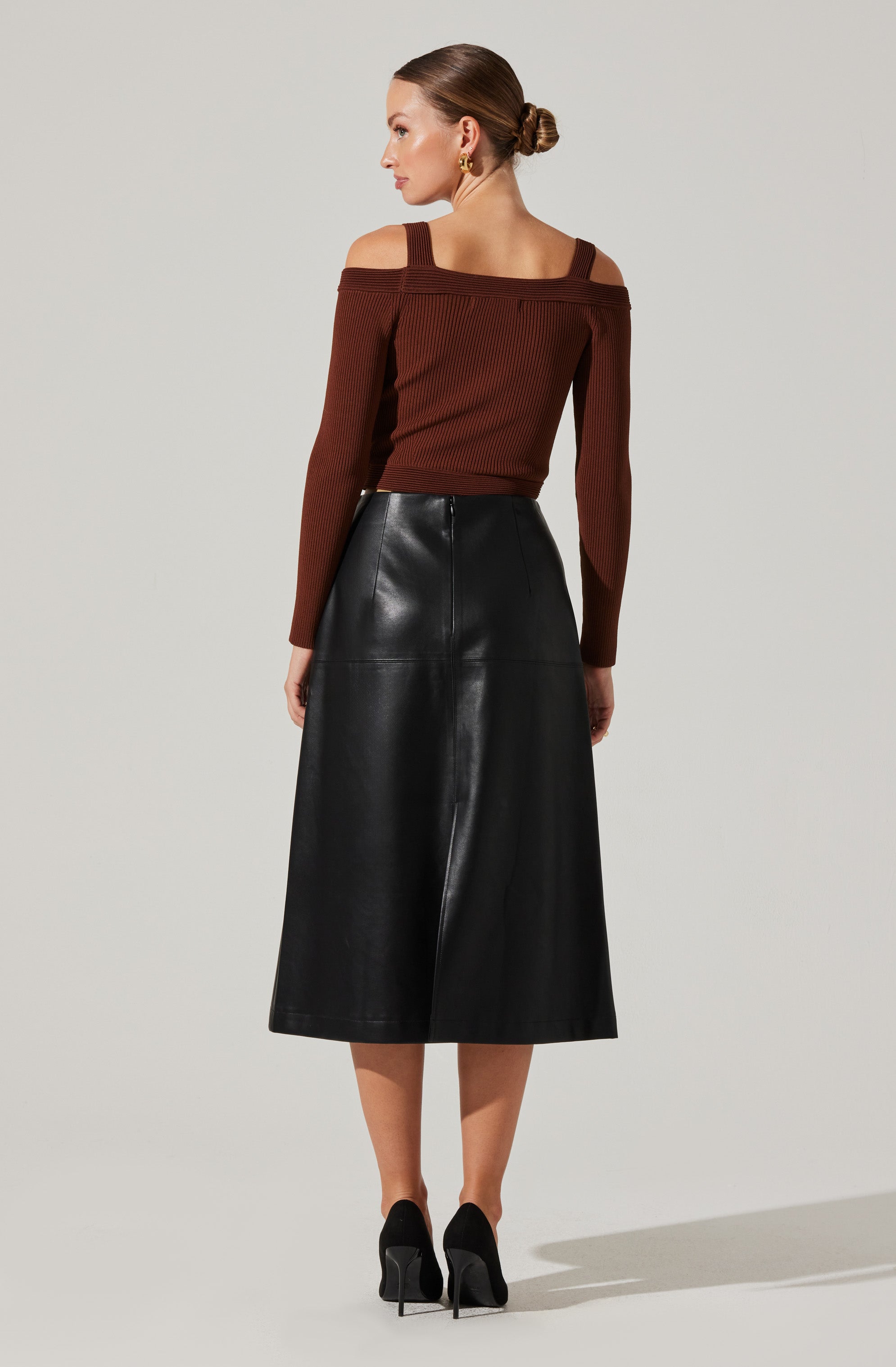 A woman is wearing a brown, ribbed, cold-shoulder sweater with wide straps and long sleeves, paired with a high-waisted, black leather A-line midi skirt. She has on black high-heeled shoes.