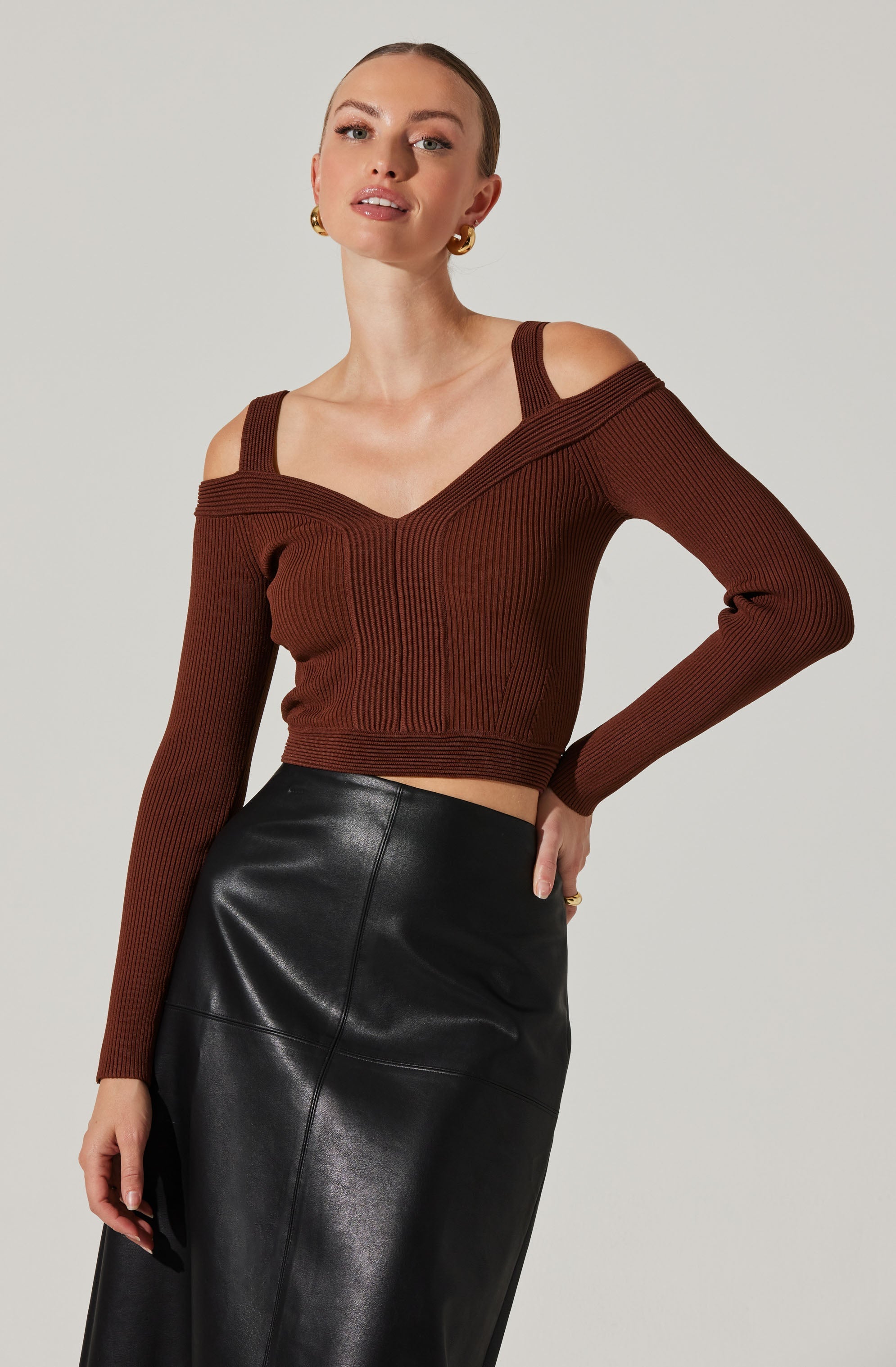 A woman is wearing a brown ribbed knit sweater with cut-out shoulders and thin straps. The sweater has a wide neckline that drapes off one shoulder, and the long sleeves are fitted. She is also wearing a high-waisted, black leather skirt. She accessorized with gold hoop earrings.