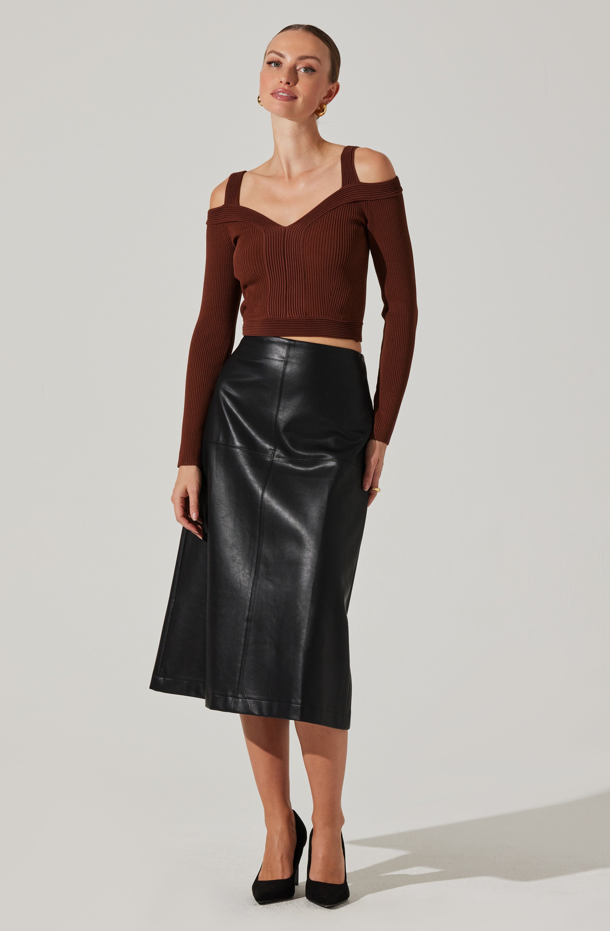A woman is wearing a dark brown, long-sleeved, off-the-shoulder knit top with shoulder cutouts and a V-neckline. She pairs it with a high-waisted, black leather midi skirt that has a slight A-line shape. She completes the outfit with black high-heeled shoes.