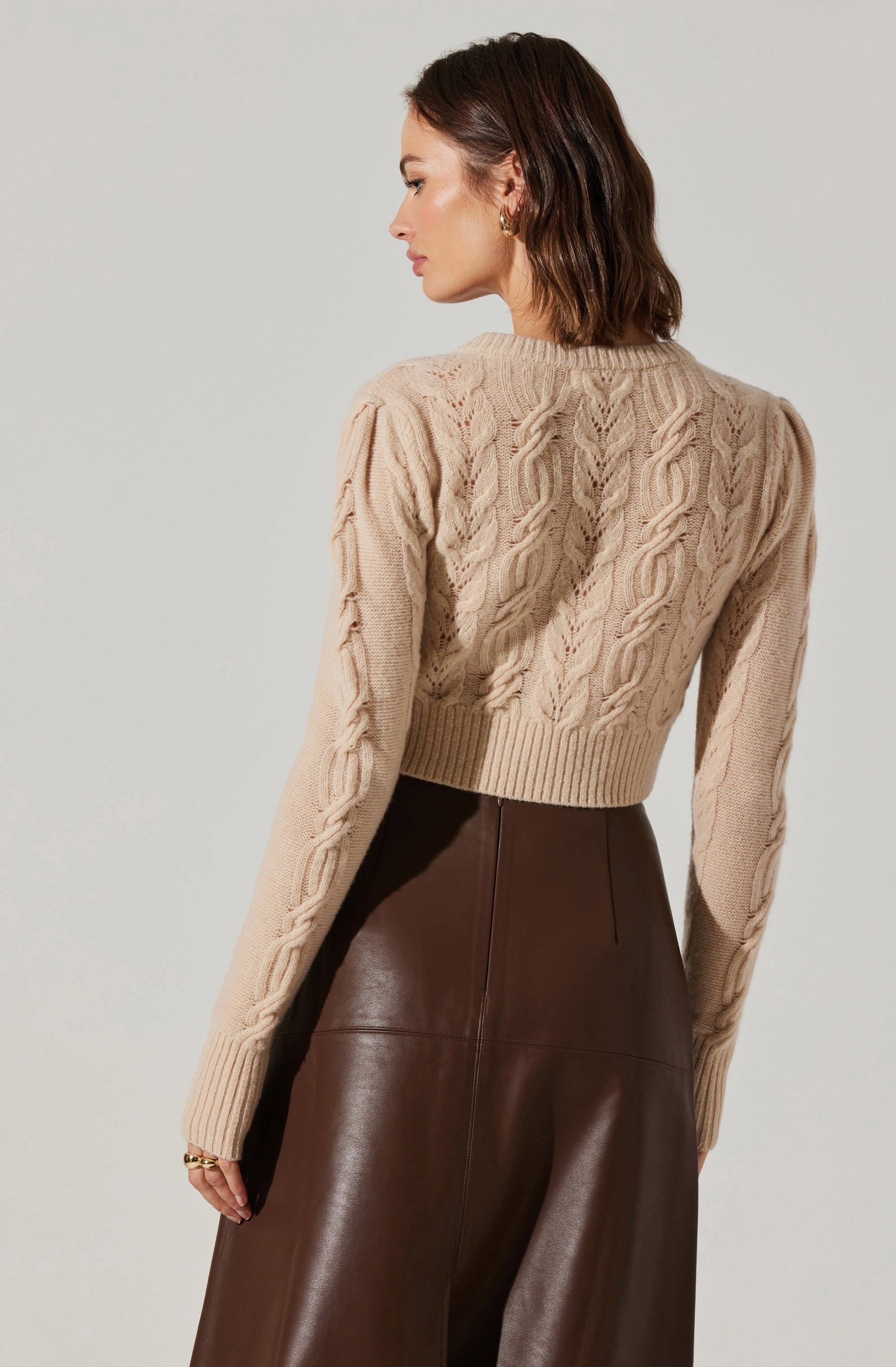 A woman is wearing a beige cable-knit sweater with intricate textured patterns and long sleeves. She pairs it with high-waisted, dark brown leather pants. The outfit is accessorized with gold hoop earrings.